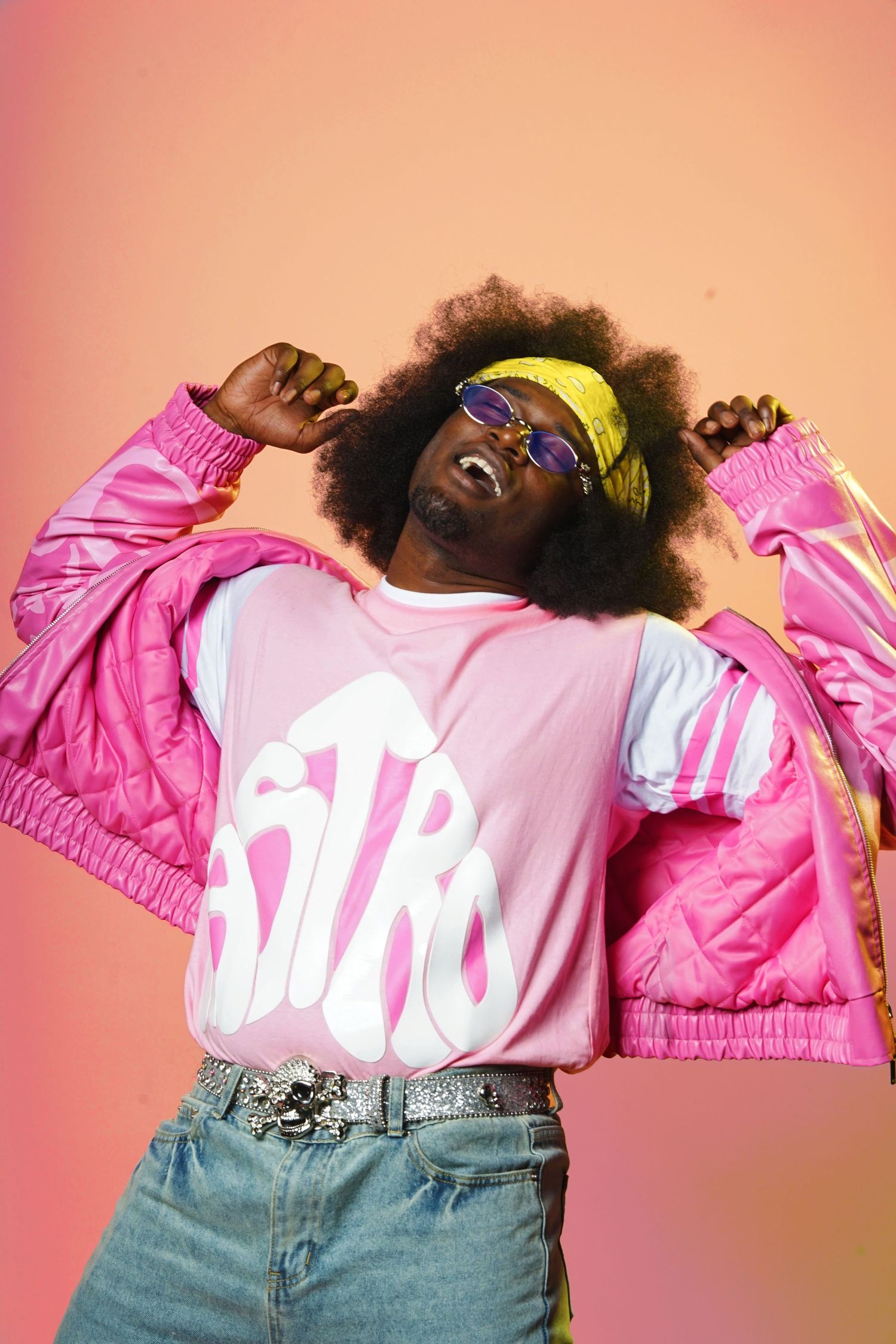 Energetic fashion shot of model in pink jacket and yellow bandana against coral background.