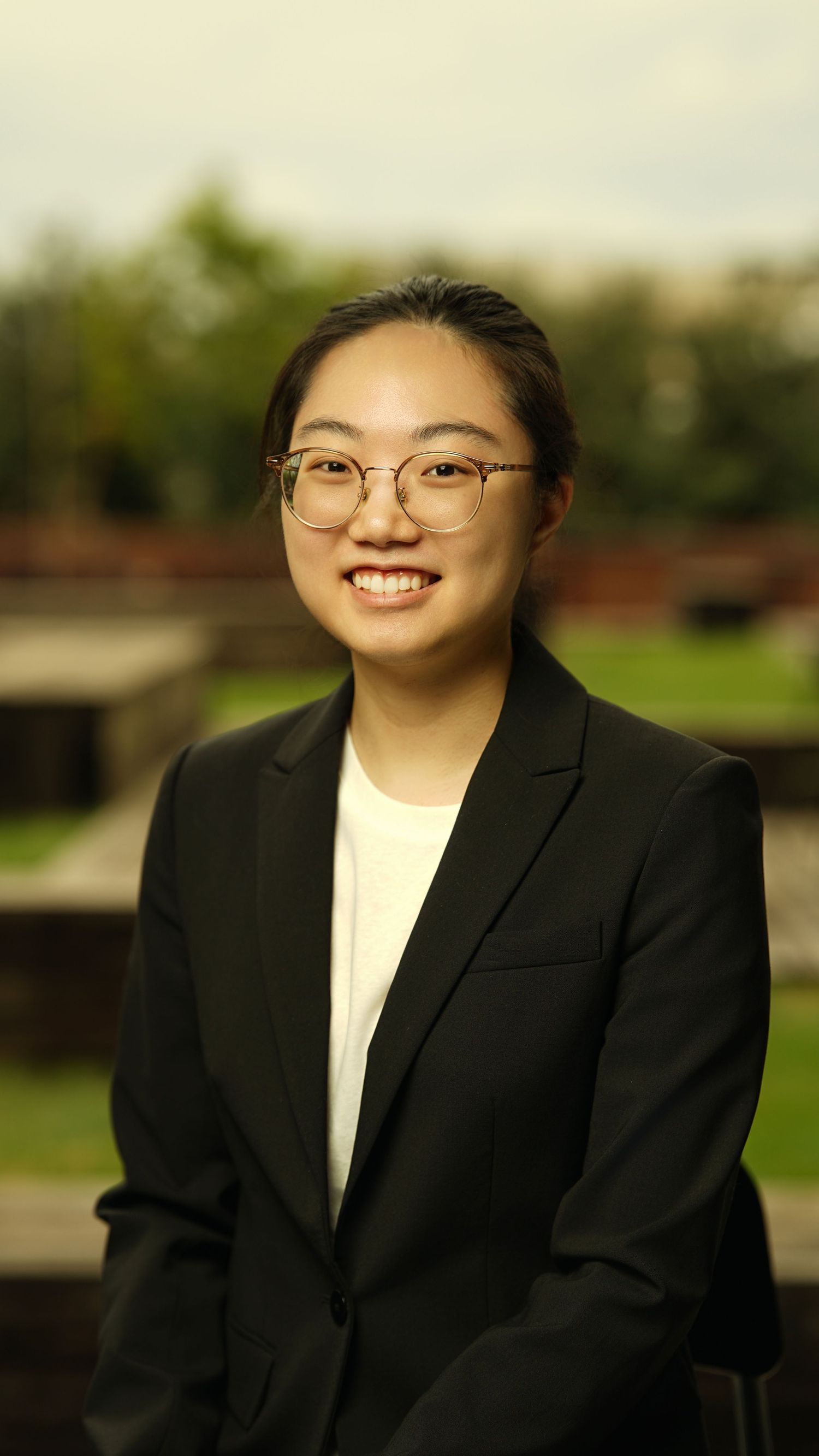 Professional in black blazer and glasses smiles warmly in outdoor portrait.