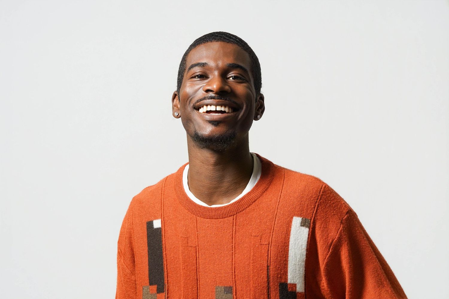 Smiling young Black man wearing an orange patterned sweater against a light gray background.