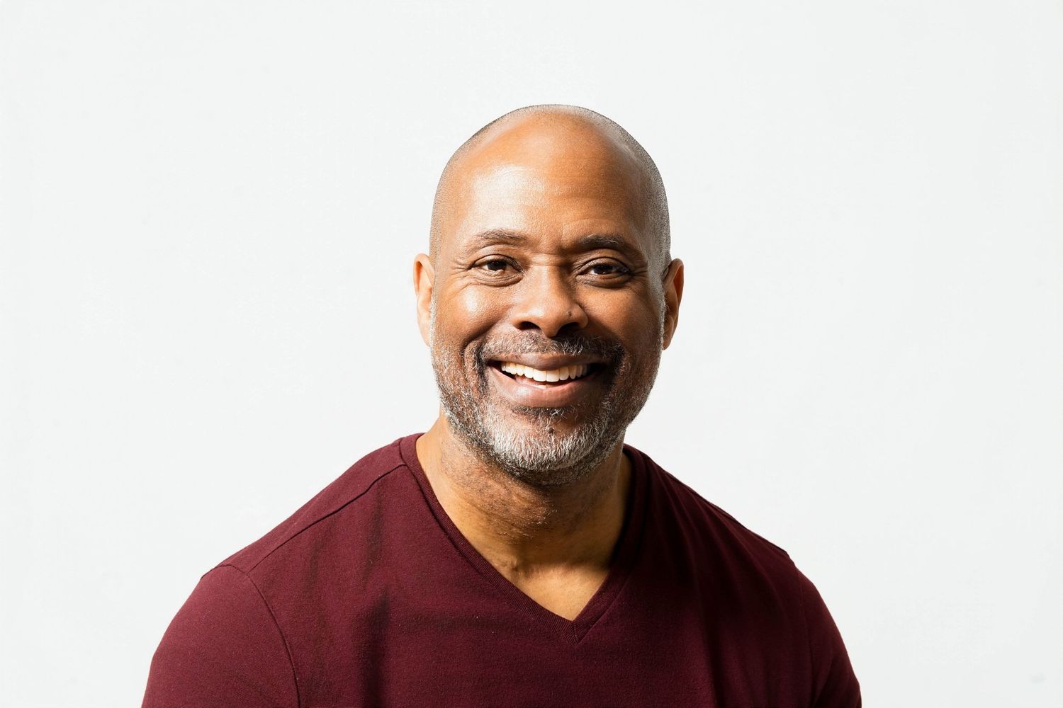 Smiling middle-aged bald Black man with gray beard wearing a dark red v-neck shirt against white background.