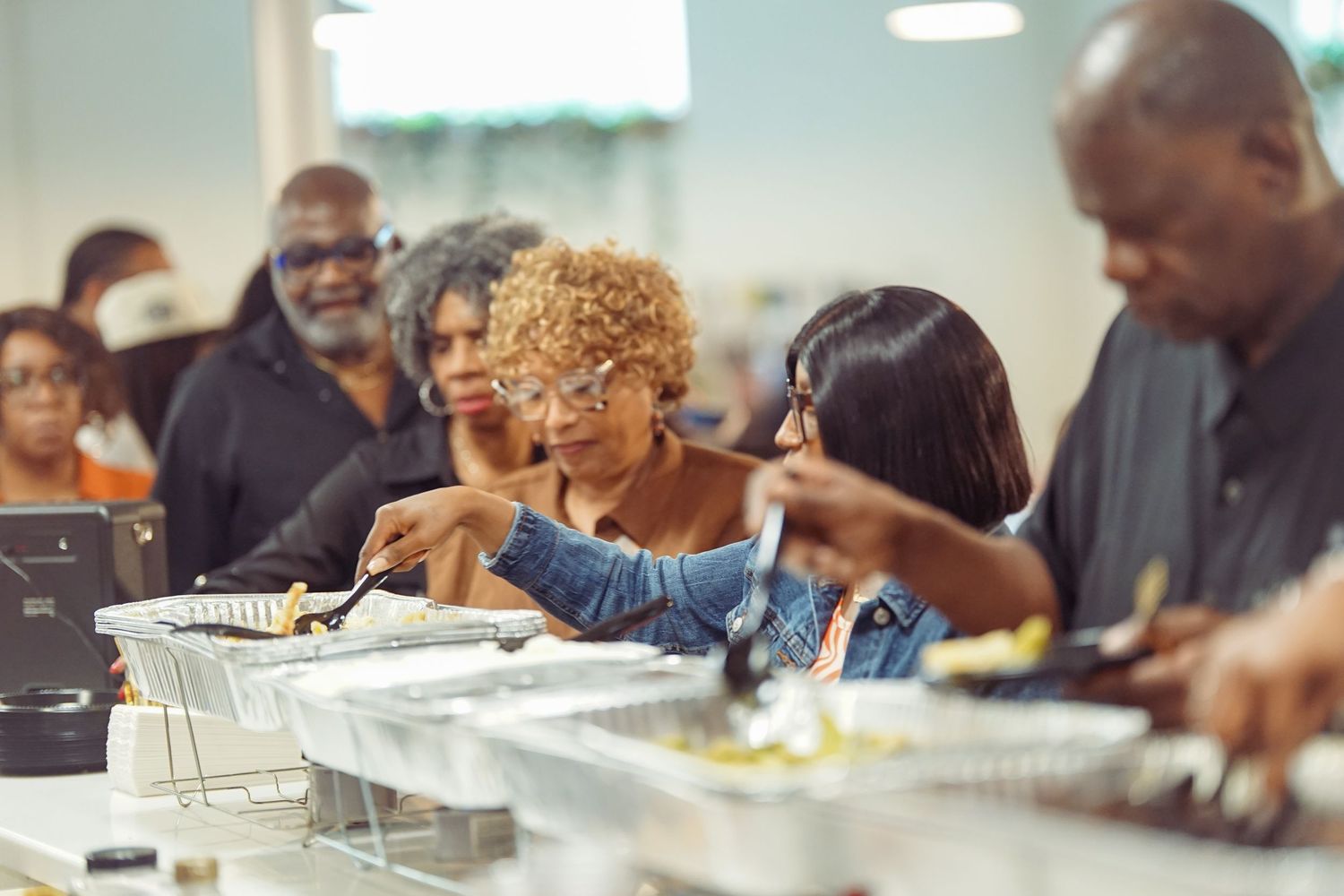 People serving themselves at a buffet line with aluminum trays of food at a community gathering event.