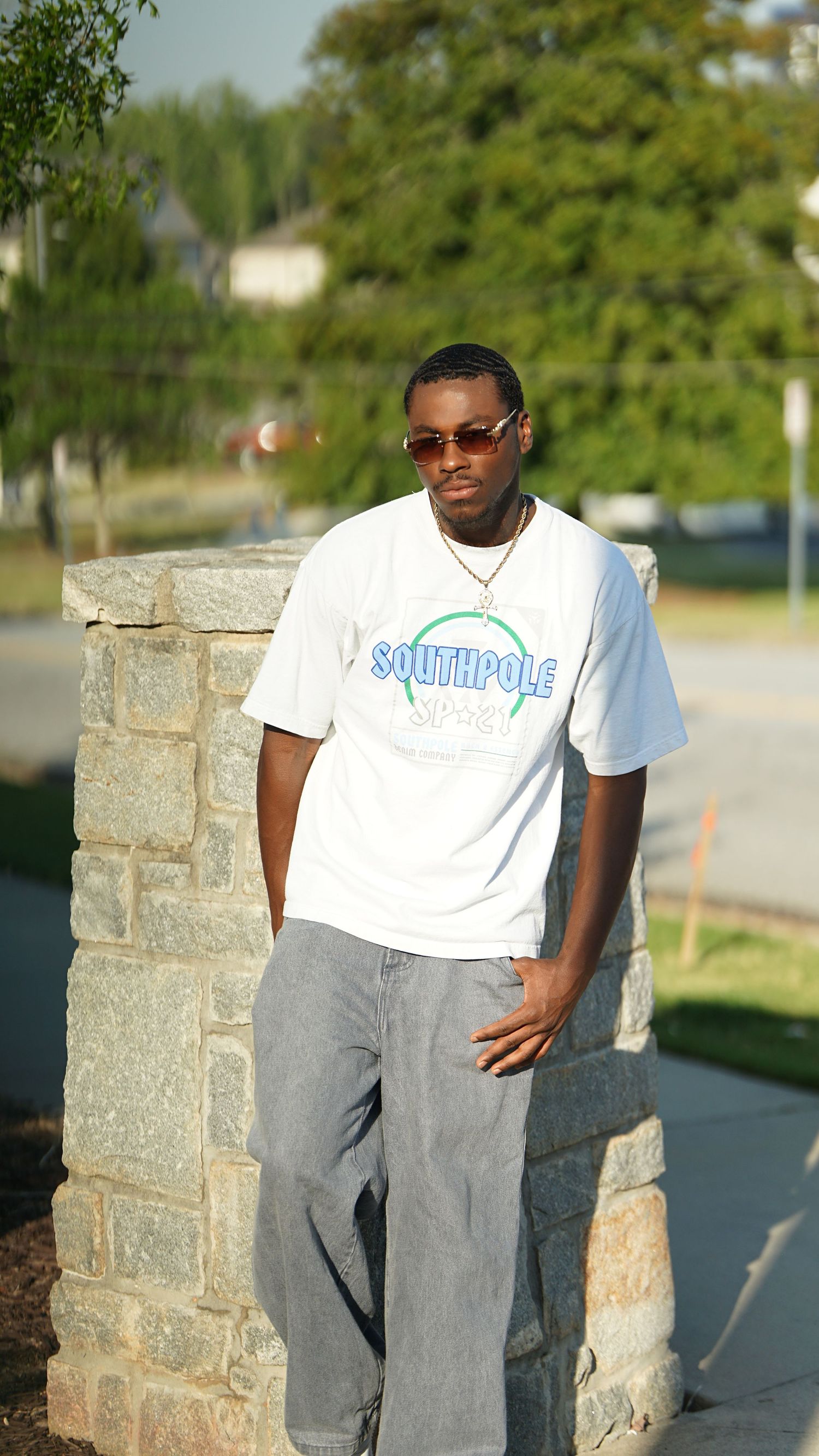 Person in white t-shirt and gray pants posing against stone wall outdoors.