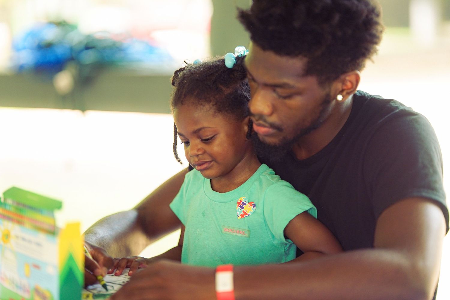 Young father and toddler daughter reading a colorful book together at an outdoor table.