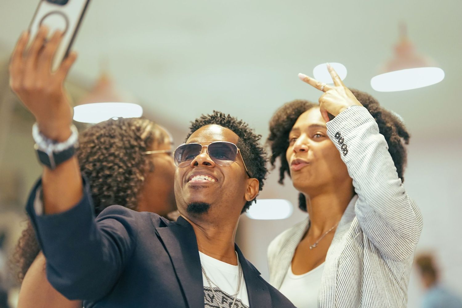 A man in sunglasses takes a selfie with a woman making a peace sign indoors.