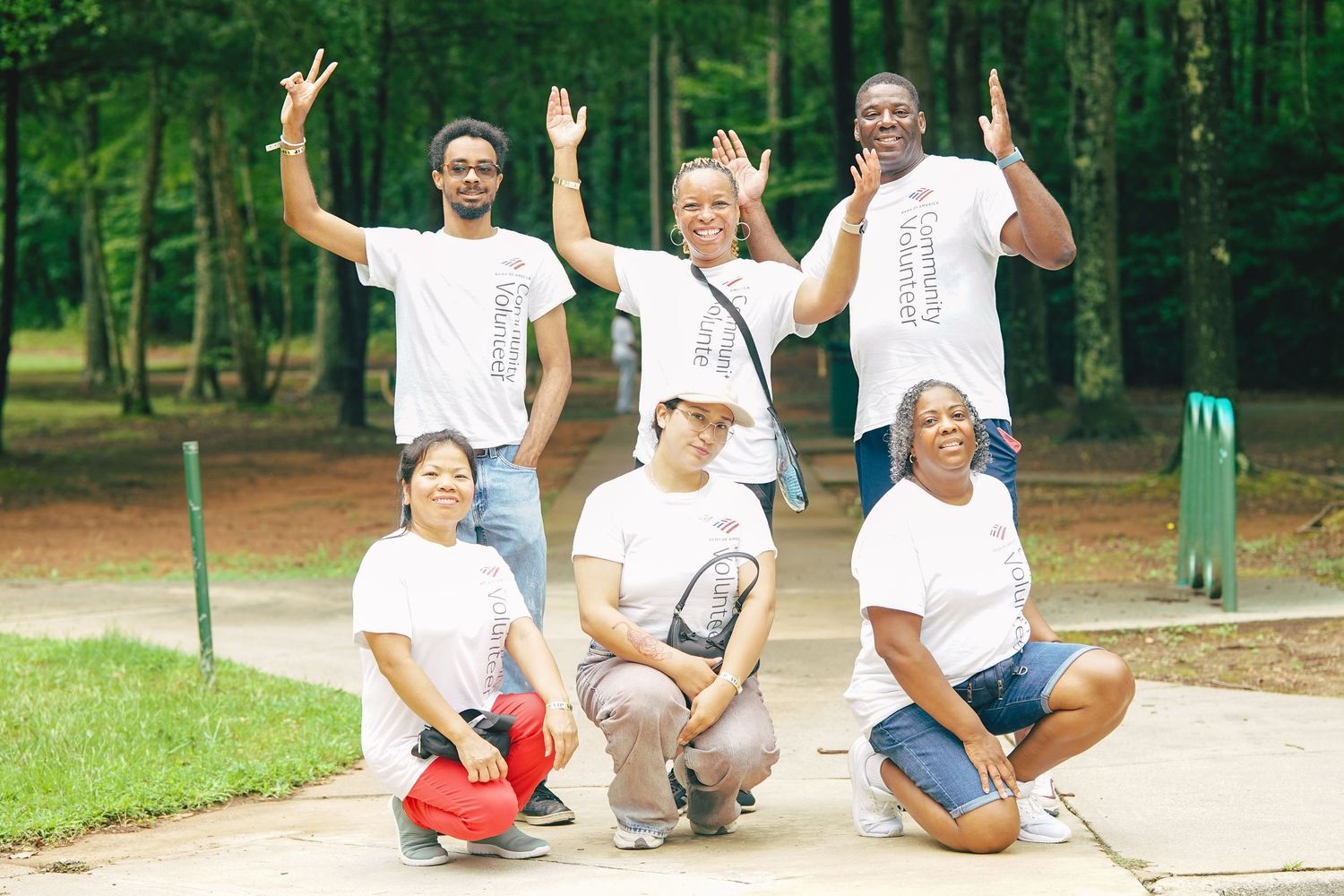 Six people in matching white t-shirts pose outdoors in a park, smiling and making peace signs for a group photo.