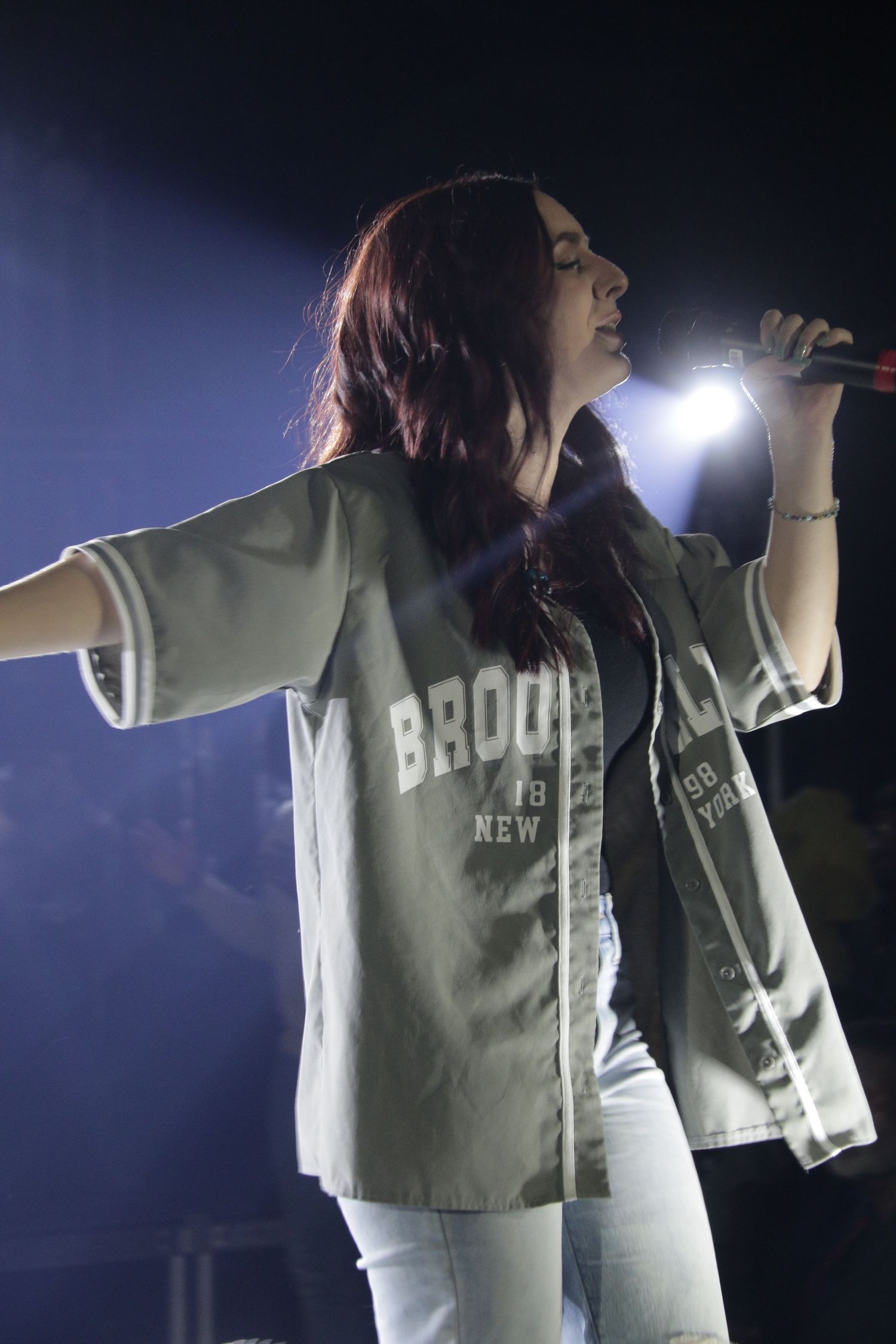 Female singer performing on stage in a Brooklyn jersey and white jeans under bright spotlights.