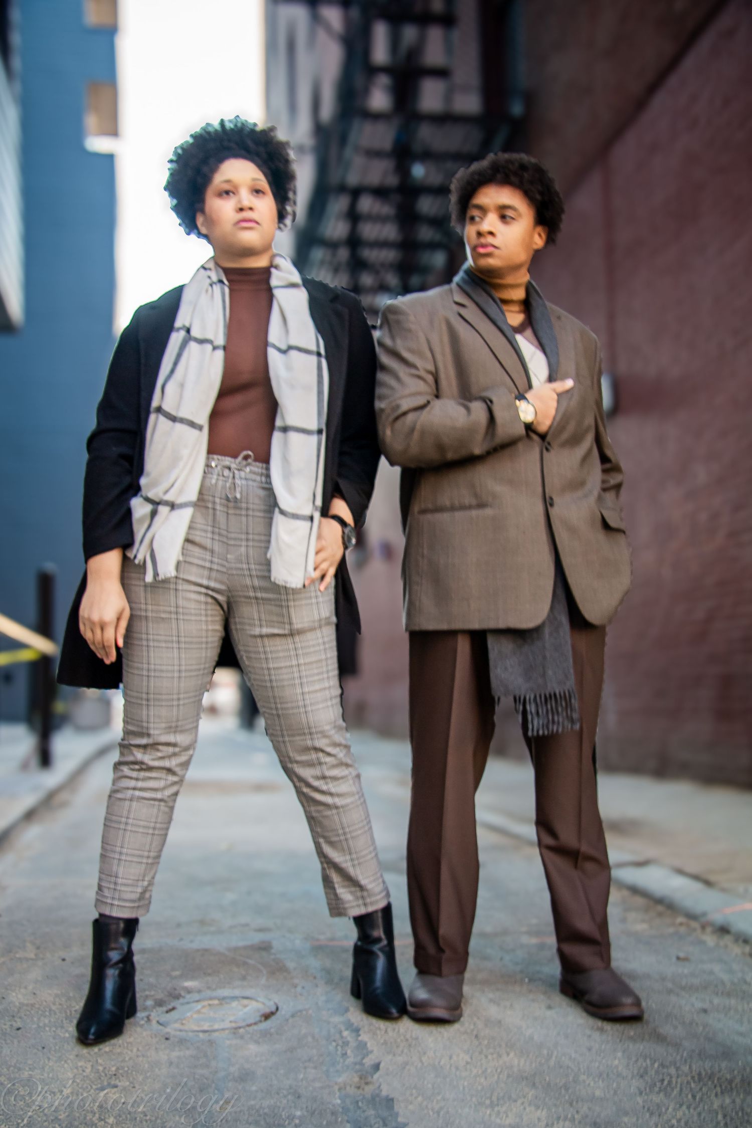 Two young Black individuals in vintage-inspired outfits pose confidently in an urban alleyway setting.