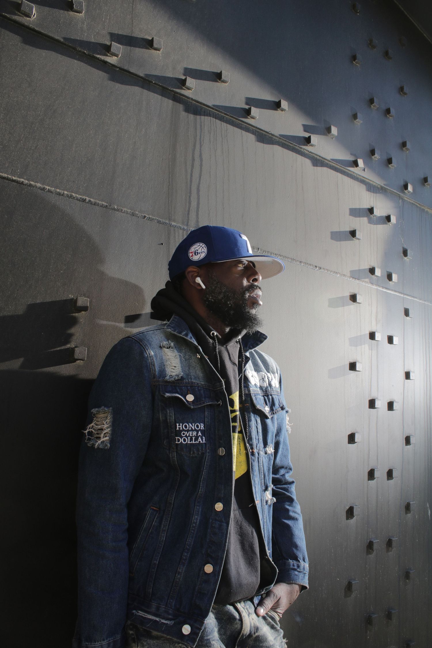 Bearded man in denim jacket and blue snapback leaning against concrete wall with earbuds.