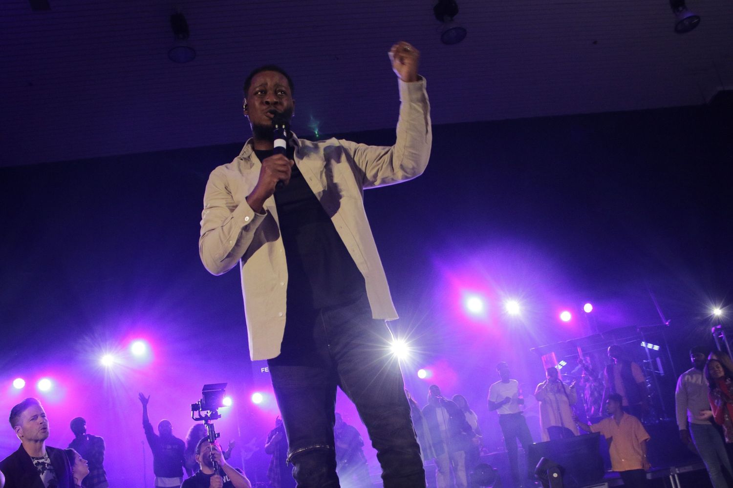 A performer in a white blazer energetically sings into a microphone on stage with purple concert lights behind him.