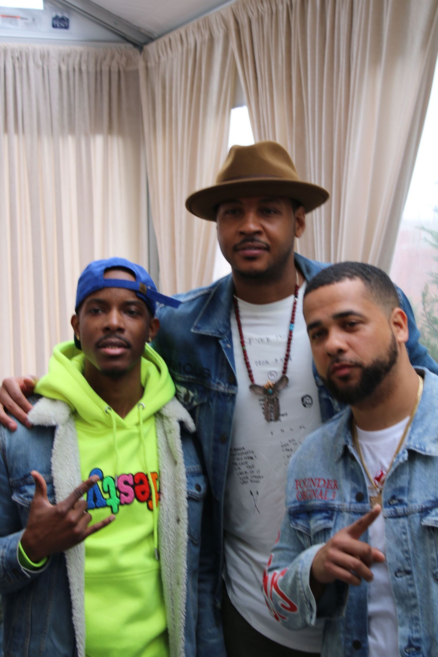 Three men pose together at an indoor event, center man wearing a tan fedora and denim jacket with accessories.