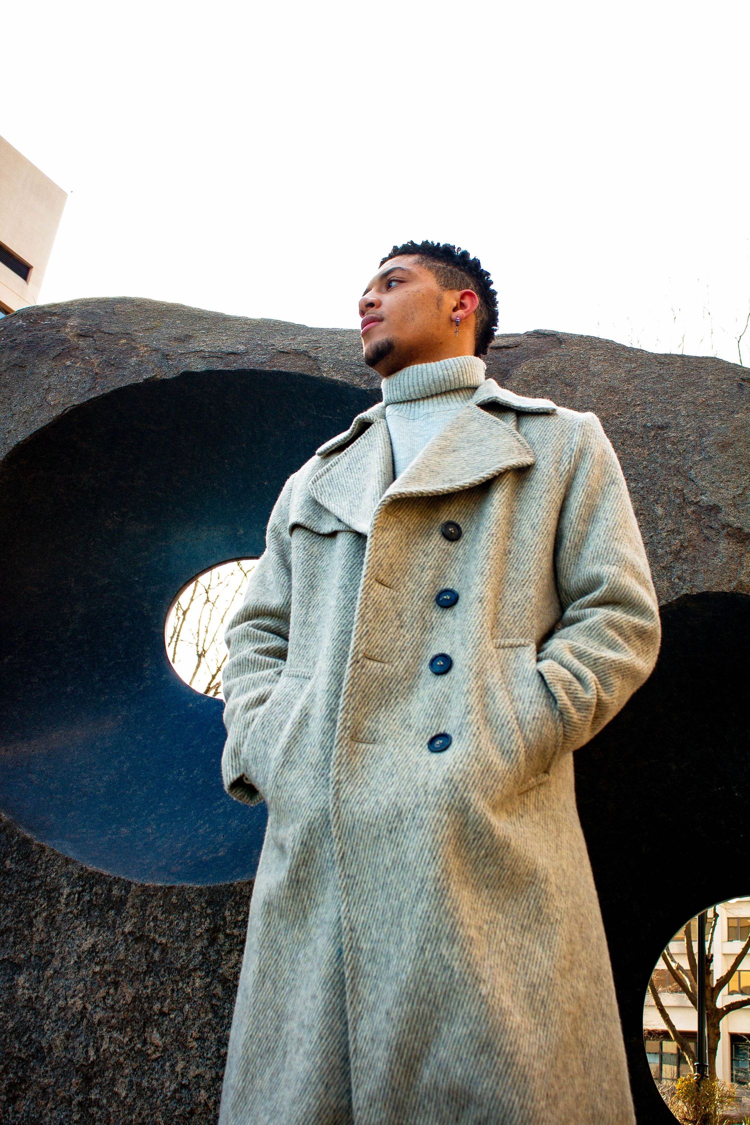 Young man in long gray overcoat standing beside large abstract stone sculpture outdoors.