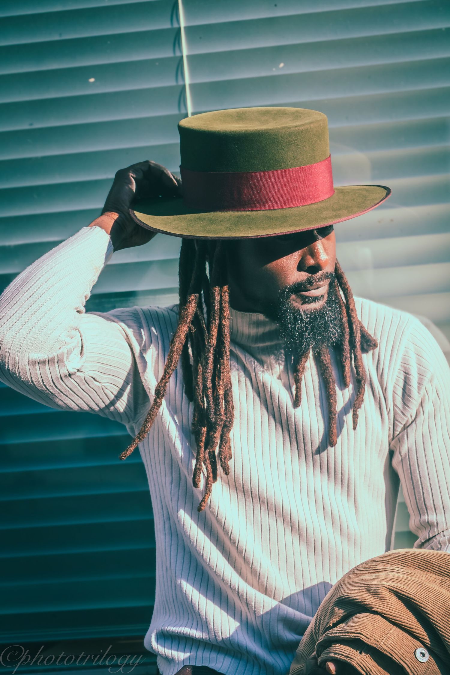 Stylish Black man with dreadlocks wearing a wide-brim hat and white ribbed sweater poses outdoors in sunlight.