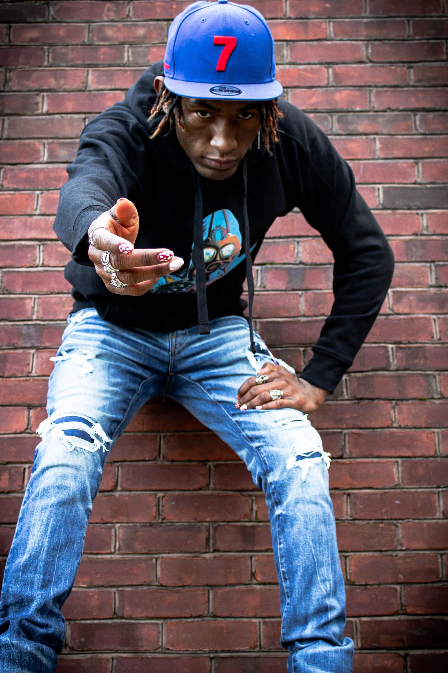 Young man in blue snapback and ripped jeans poses against brick wall.