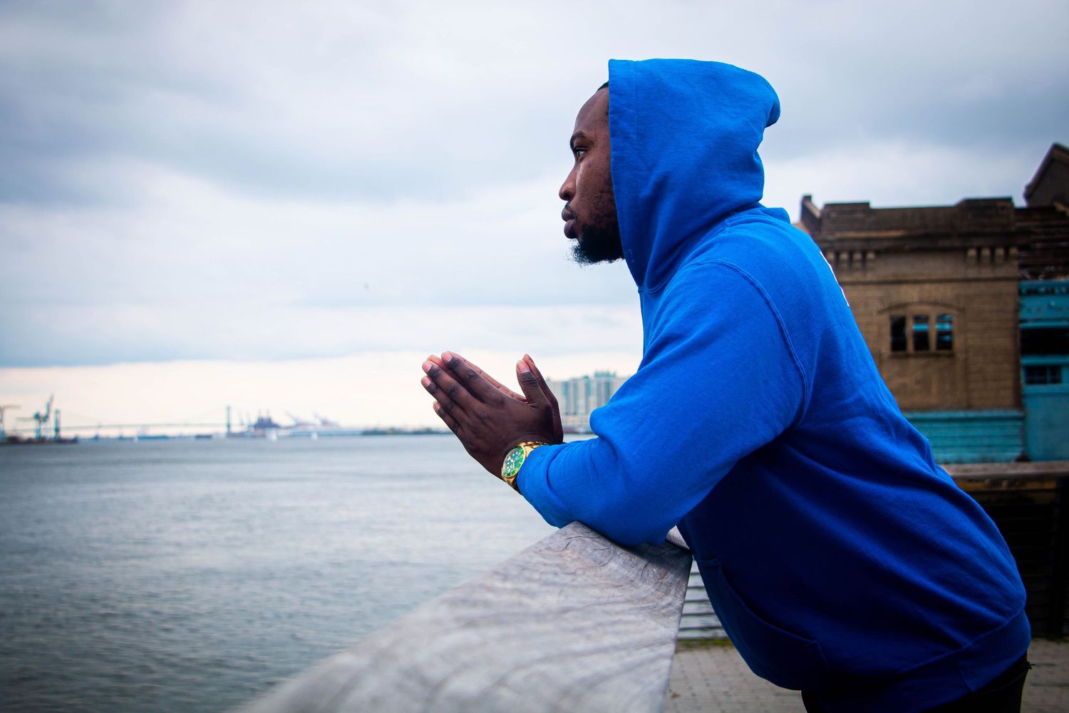 Man in blue hoodie leans on railing overlooking waterfront on cloudy day.