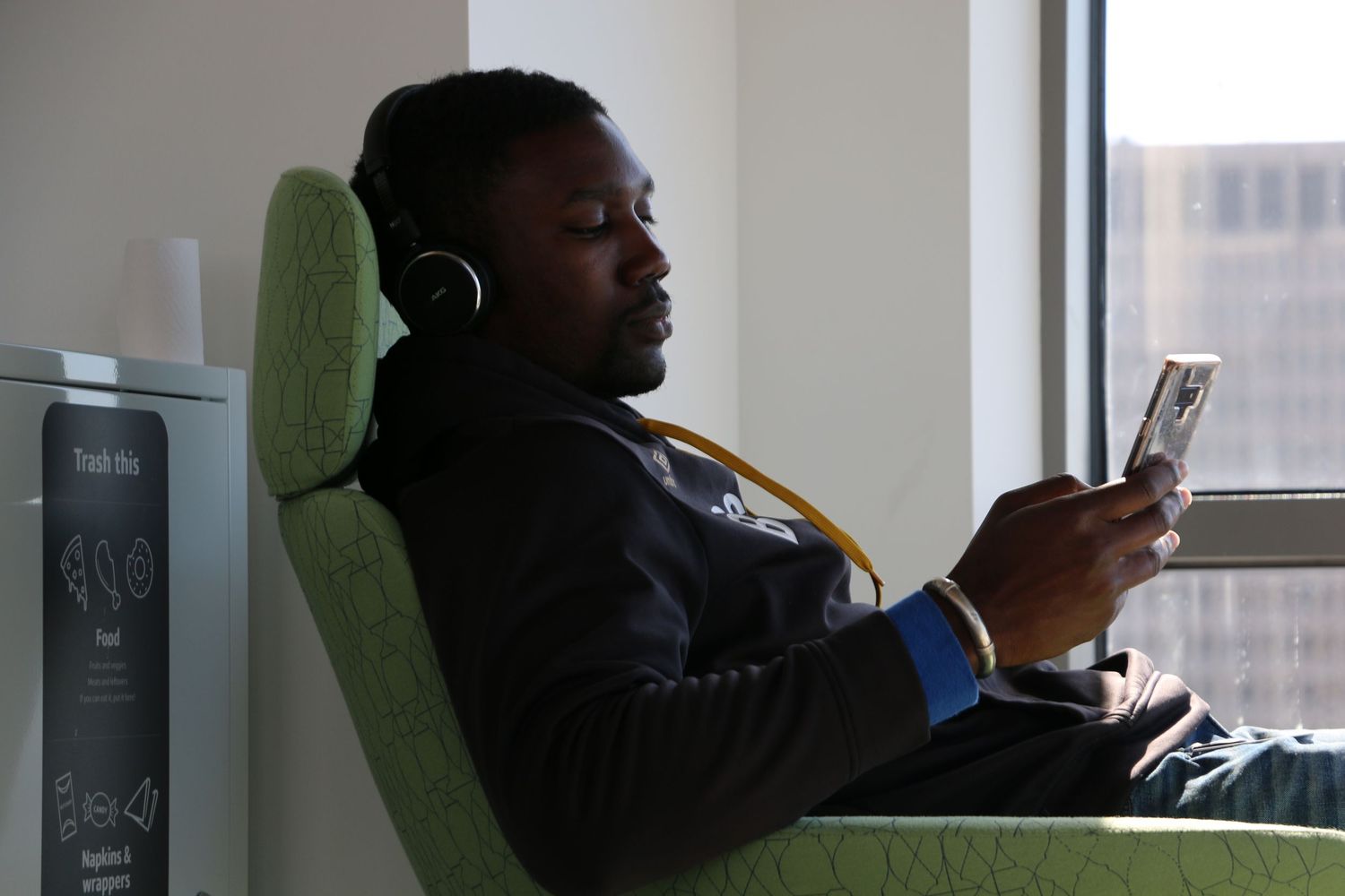 A man wearing headphones relaxes in a green chair while looking at his smartphone near a window.