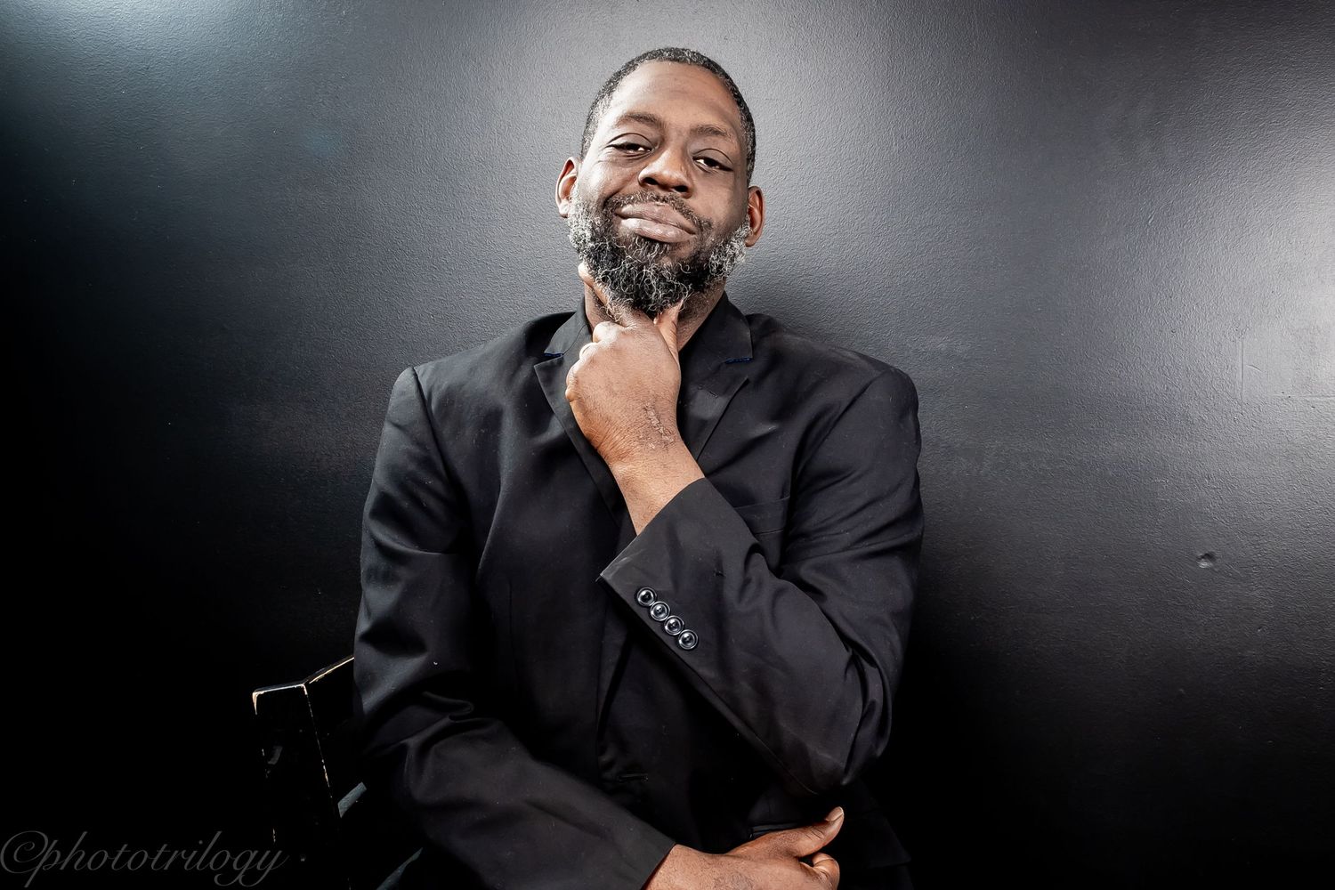 A well-dressed man in a black suit smiles warmly while posing against a dark studio background.