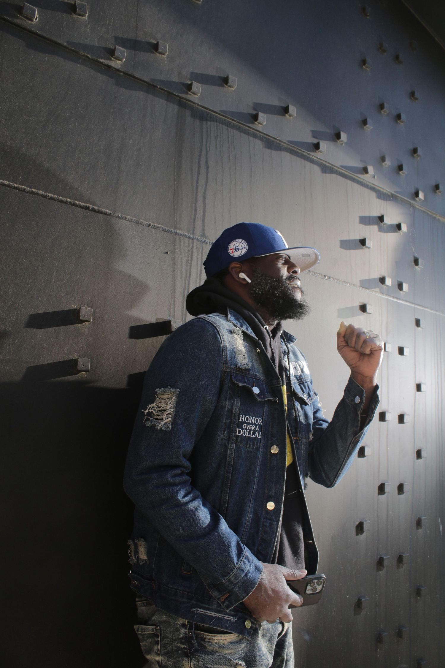 Bearded Black man in denim jacket and blue cap leaning against a textured metal wall raising his fist.