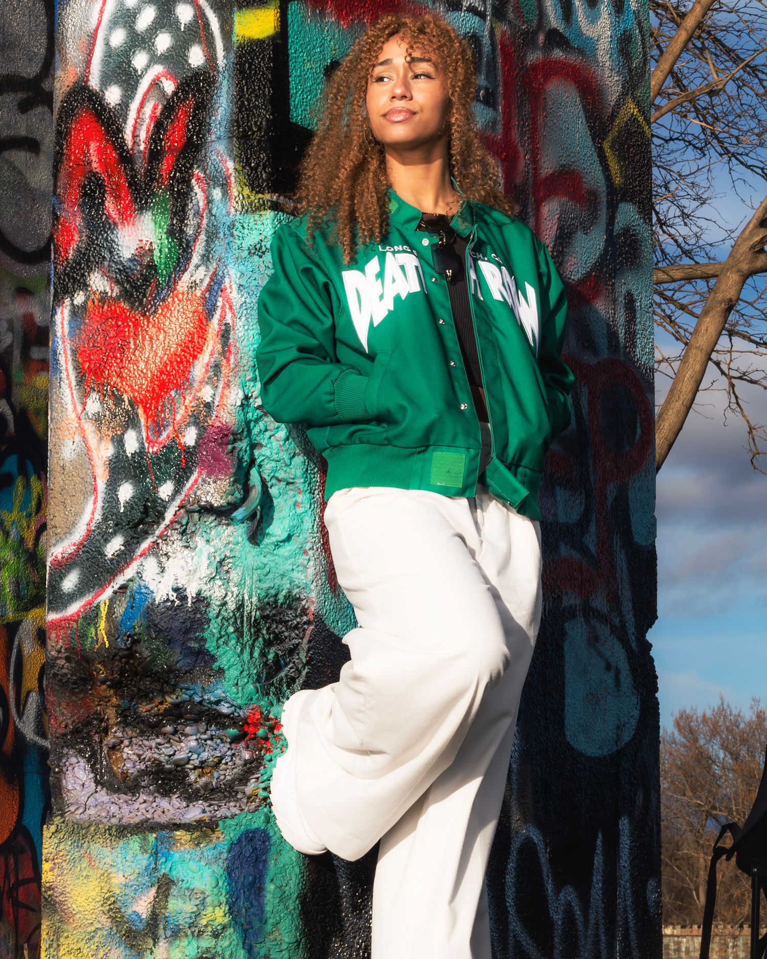 A young woman in a green jacket and white pants poses confidently against a colorful graffiti wall outdoors.