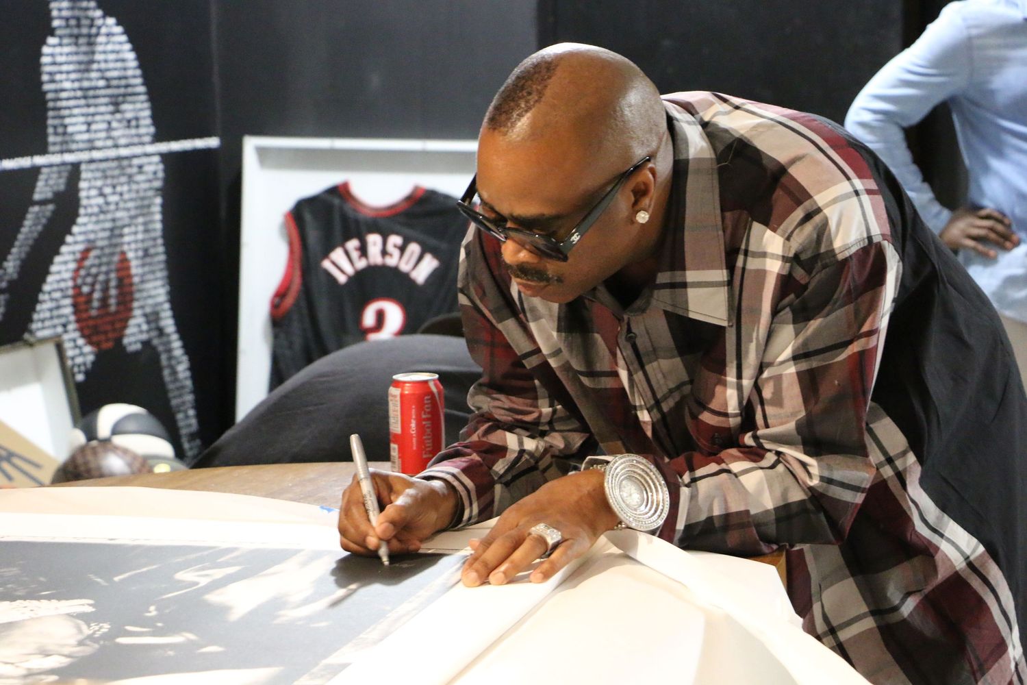 Black man in plaid shirt and sunglasses signs autographs at an event with basketball artwork in background.