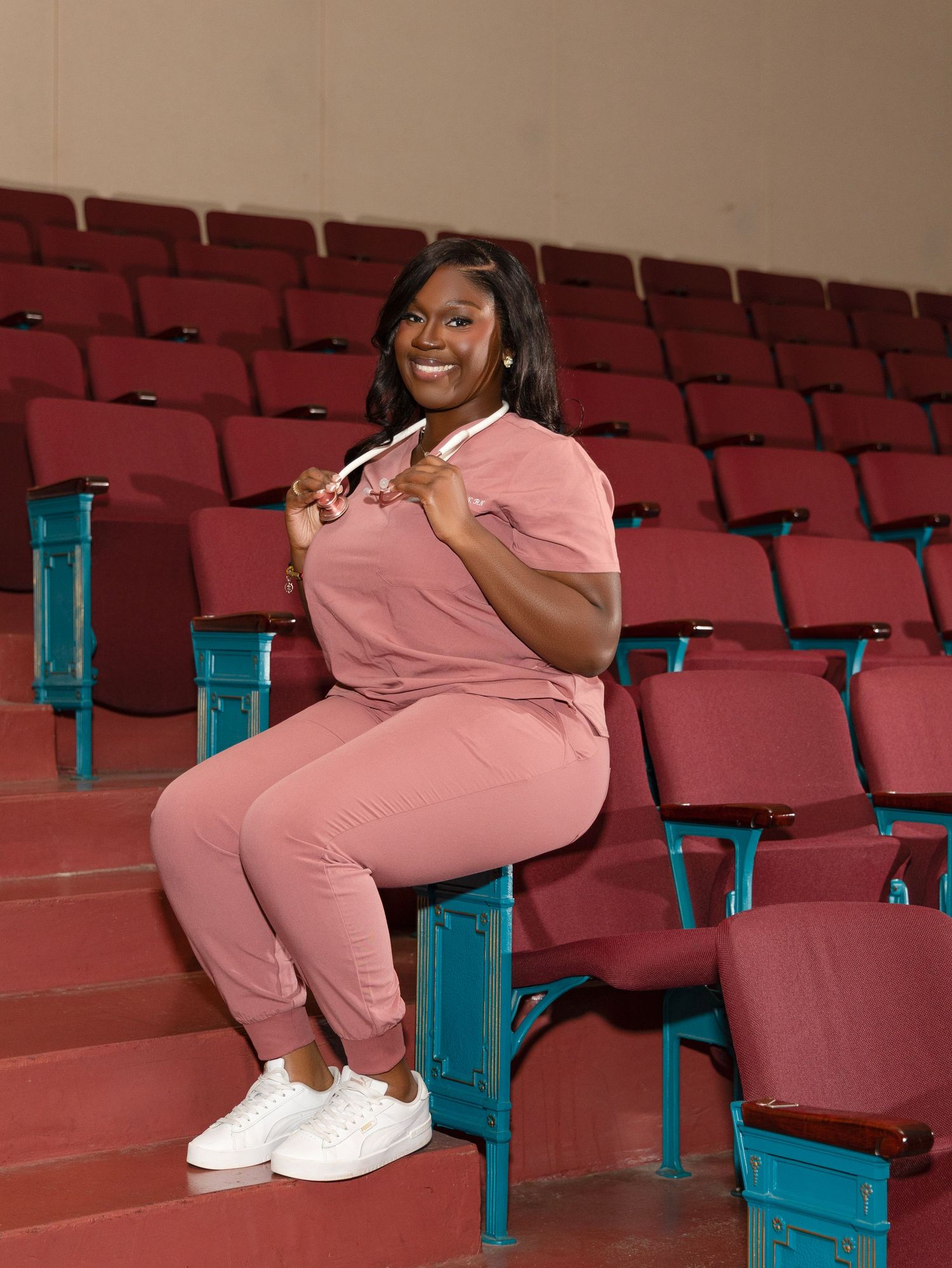 A person wearing a pink jumpsuit sits smiling in turquoise theater seats with white sneakers.