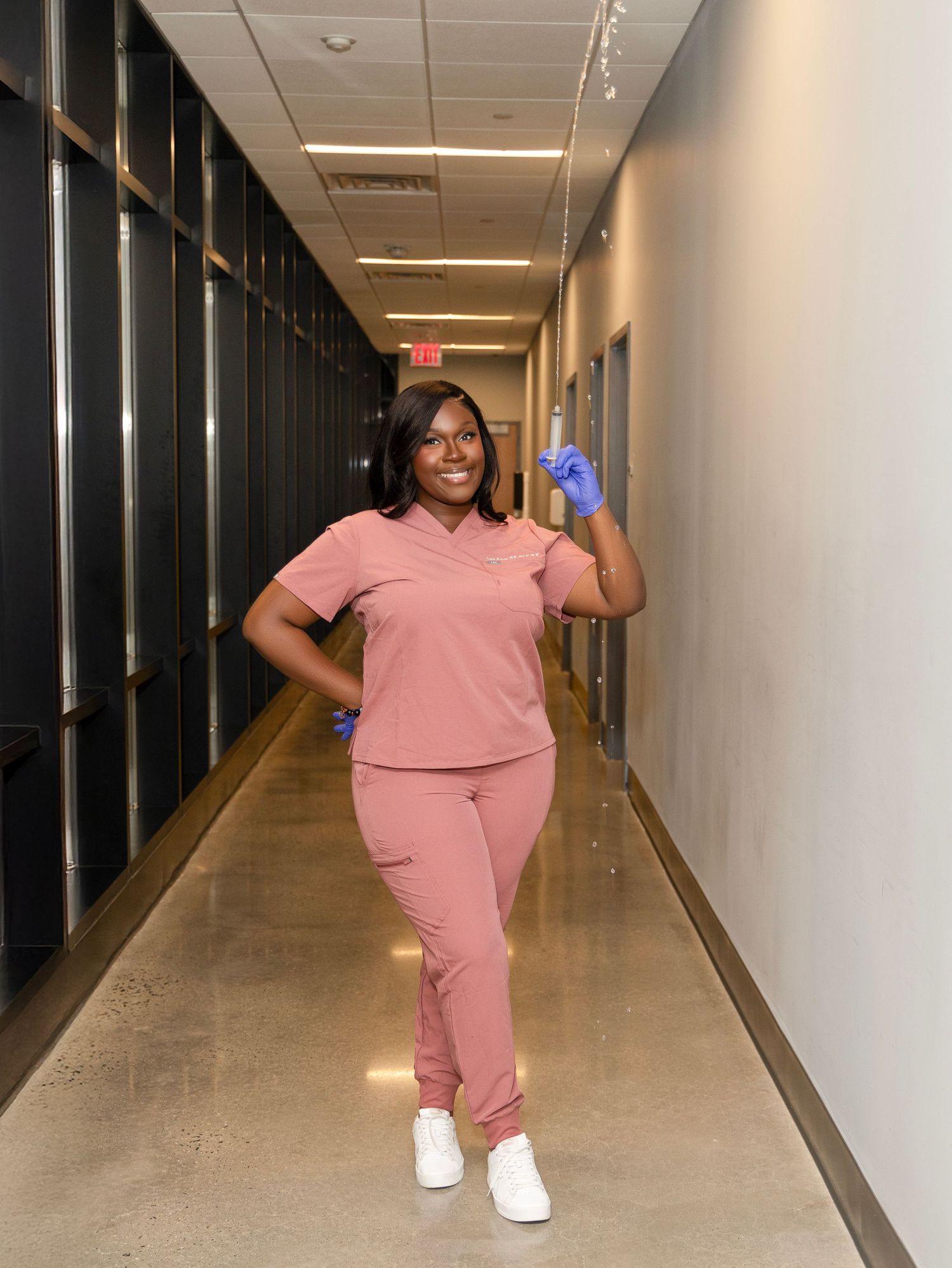 Healthcare worker in pink scrubs and white shoes stands posing in a long hospital hallway.