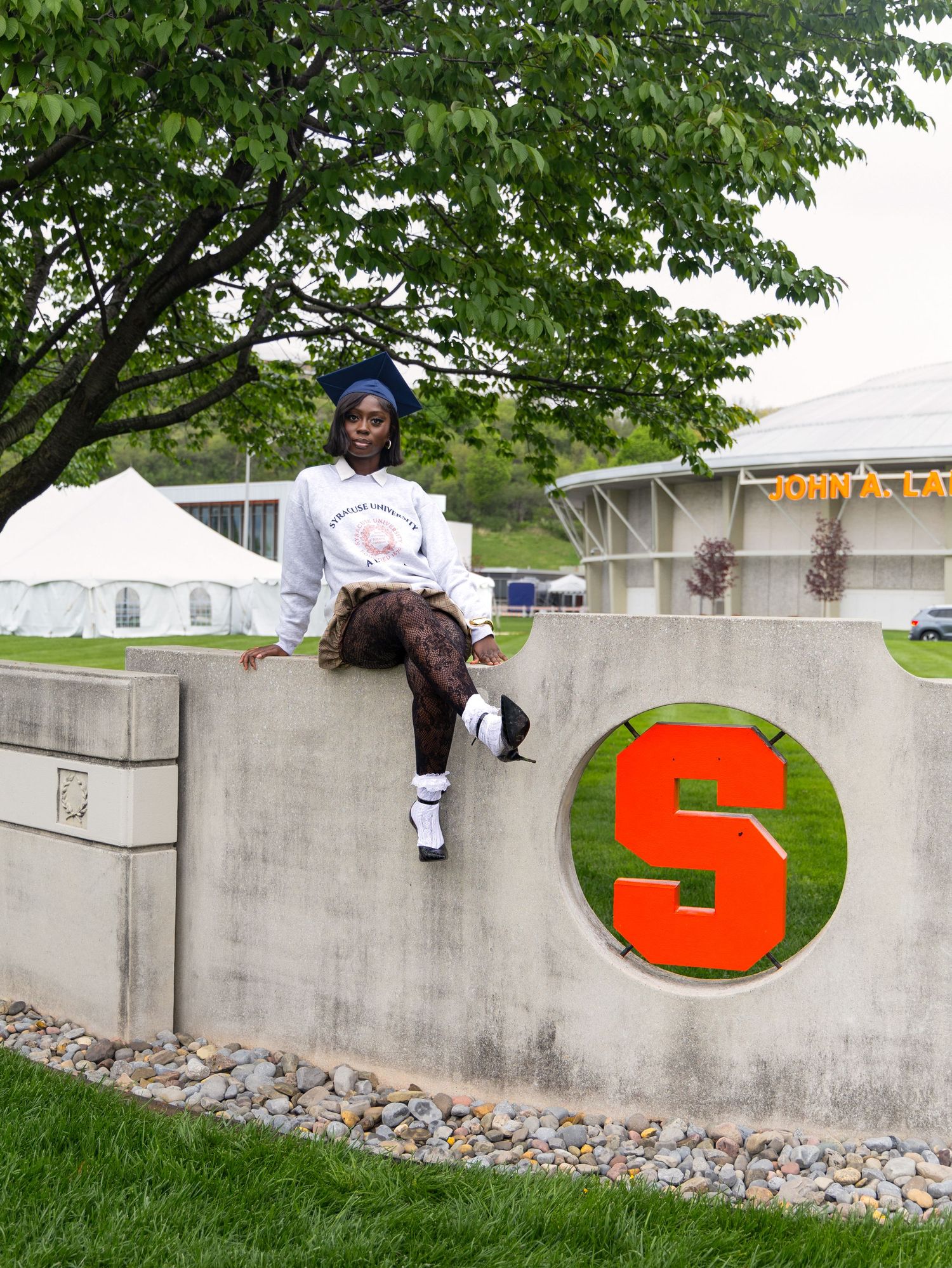 Someone sits casually on a concrete wall with a red 'S' logo, surrounded by green trees and white tents in the background.