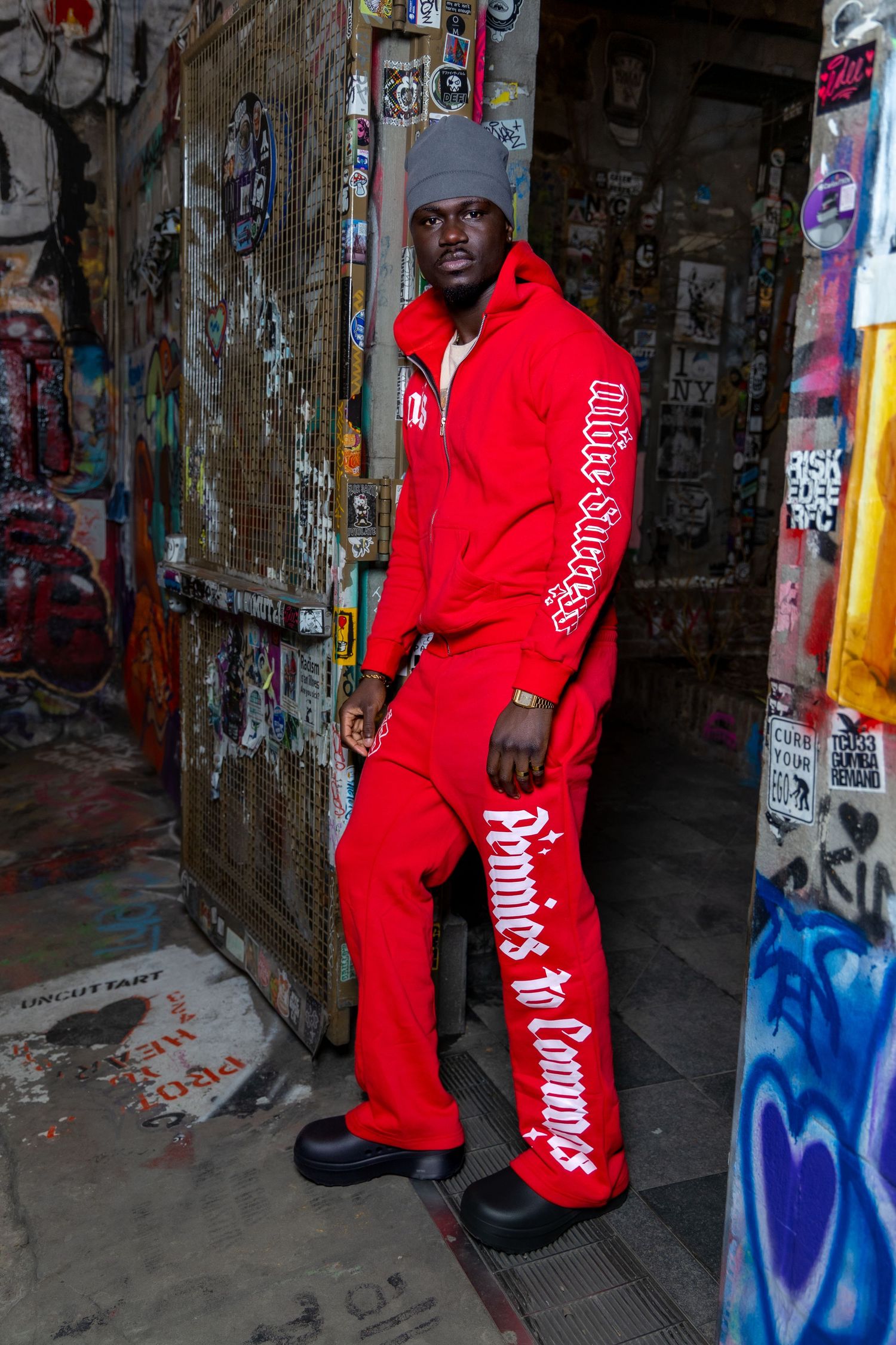 Person wearing bright red sweatsuit and beanie poses in graffiti-covered urban alleyway.