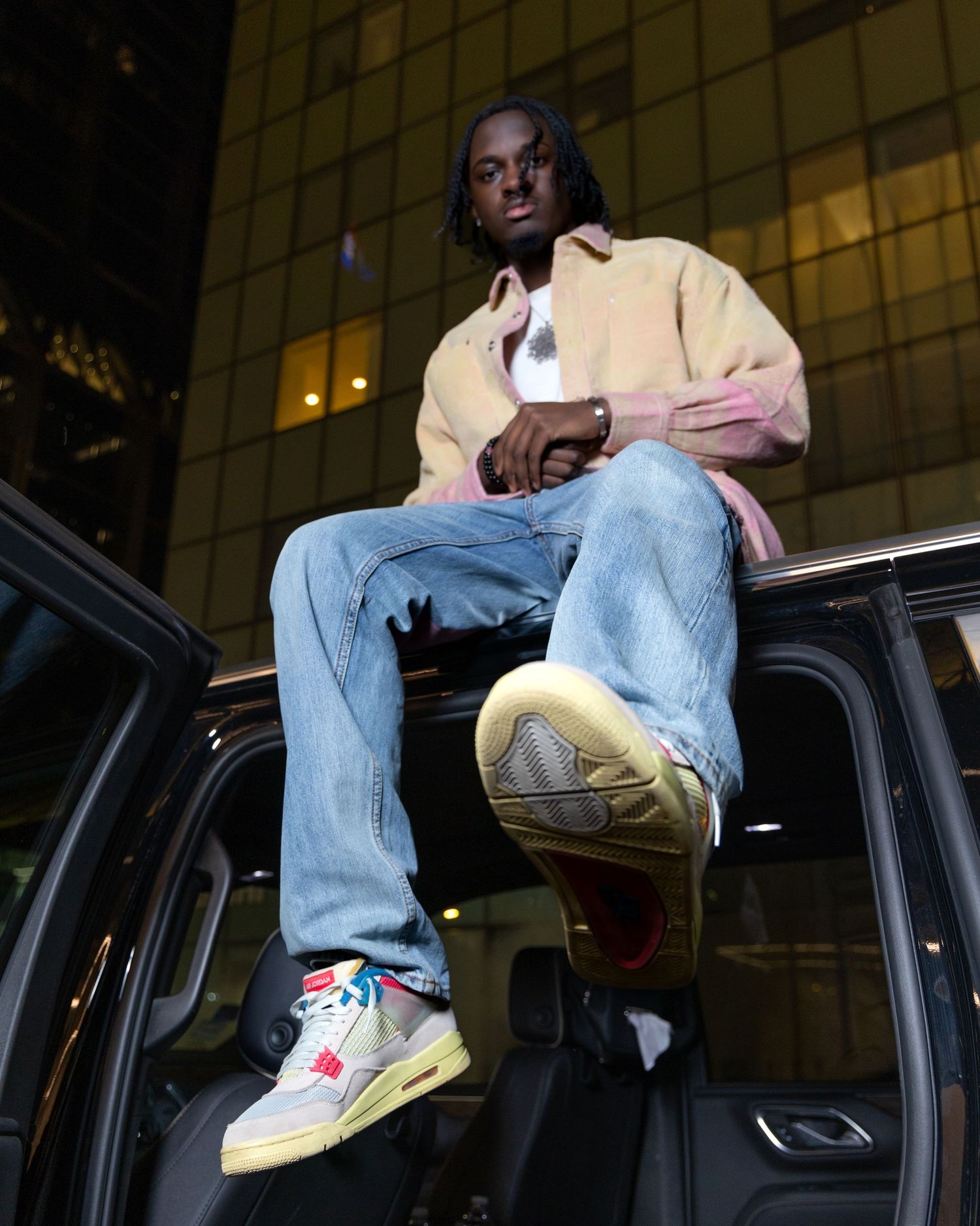 Stylish model sitting on car door wearing light jacket and jeans with sneakers against urban building backdrop.