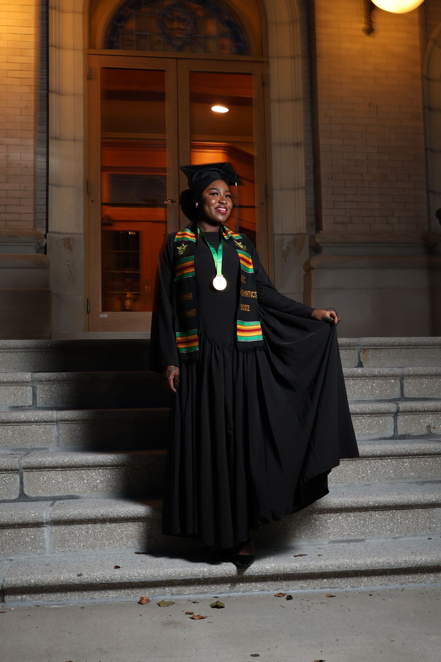 Graduate in black academic gown with kente stole poses on historic building steps at night.