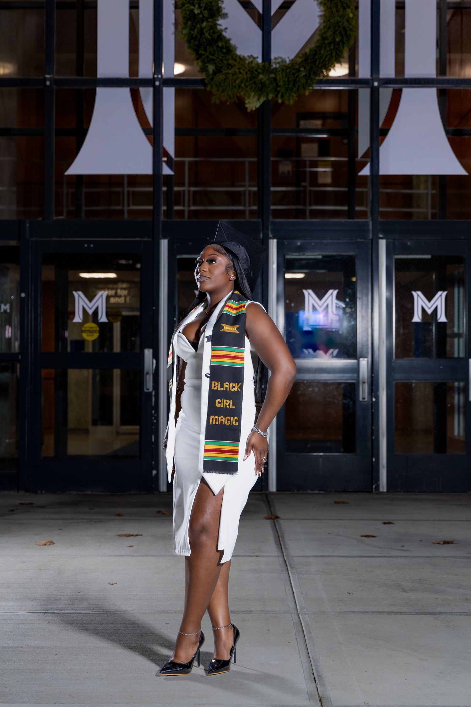 Graduate in white dress and kente stole poses confidently outside building with wreath.
