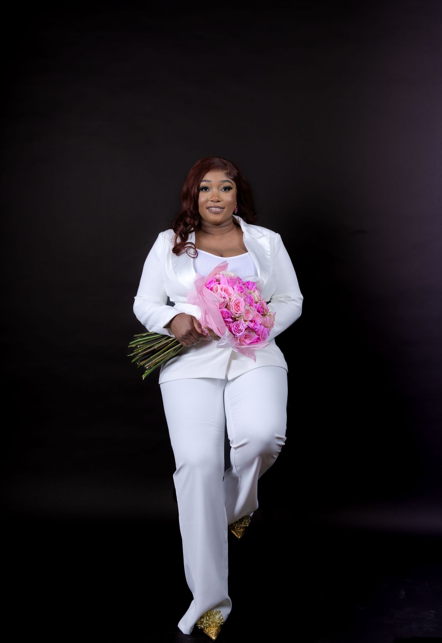 Model in all-white ensemble holding pink flowers against dark background.
