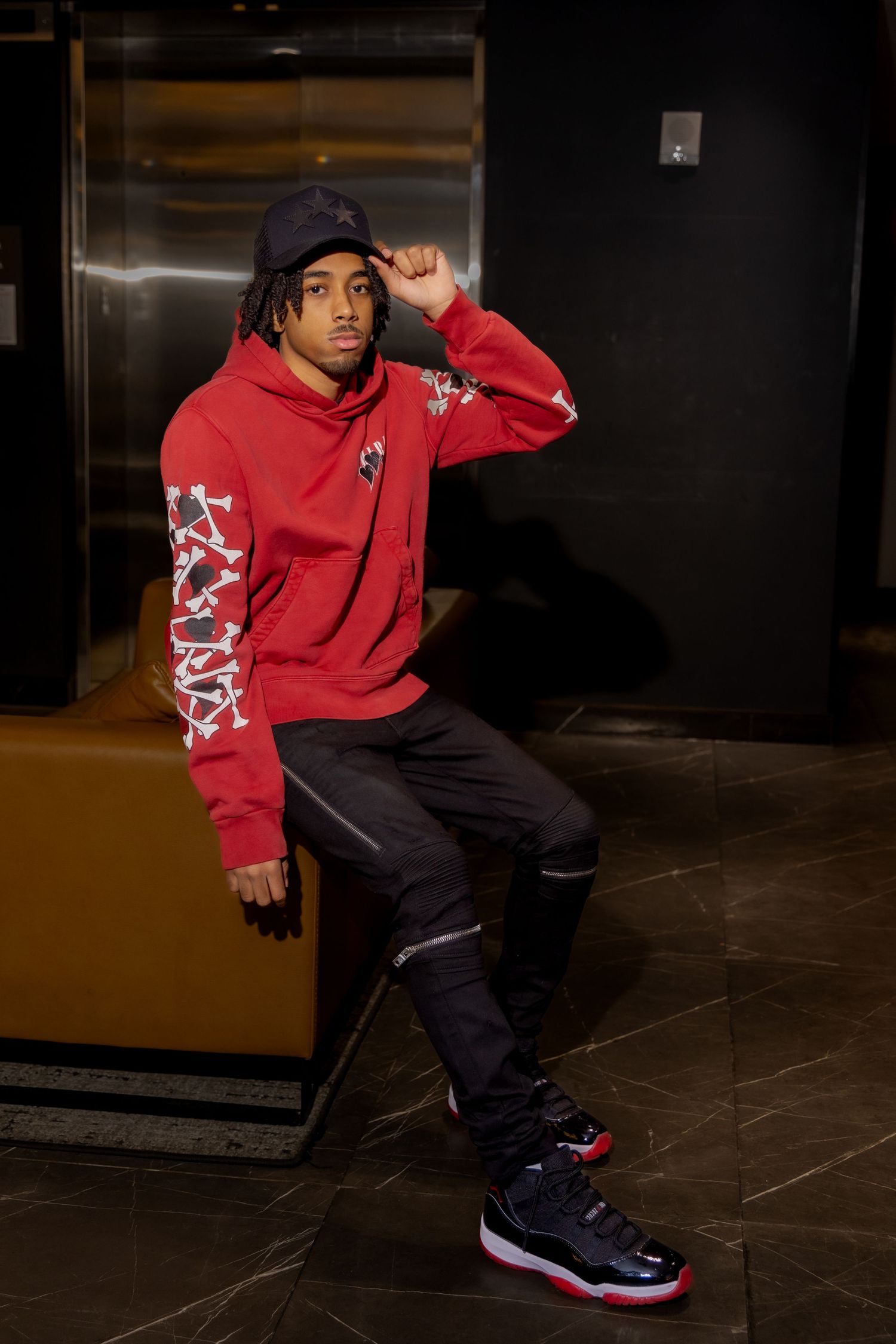 Model in red hoodie and dark pants posing against elevator with Jordan sneakers.
