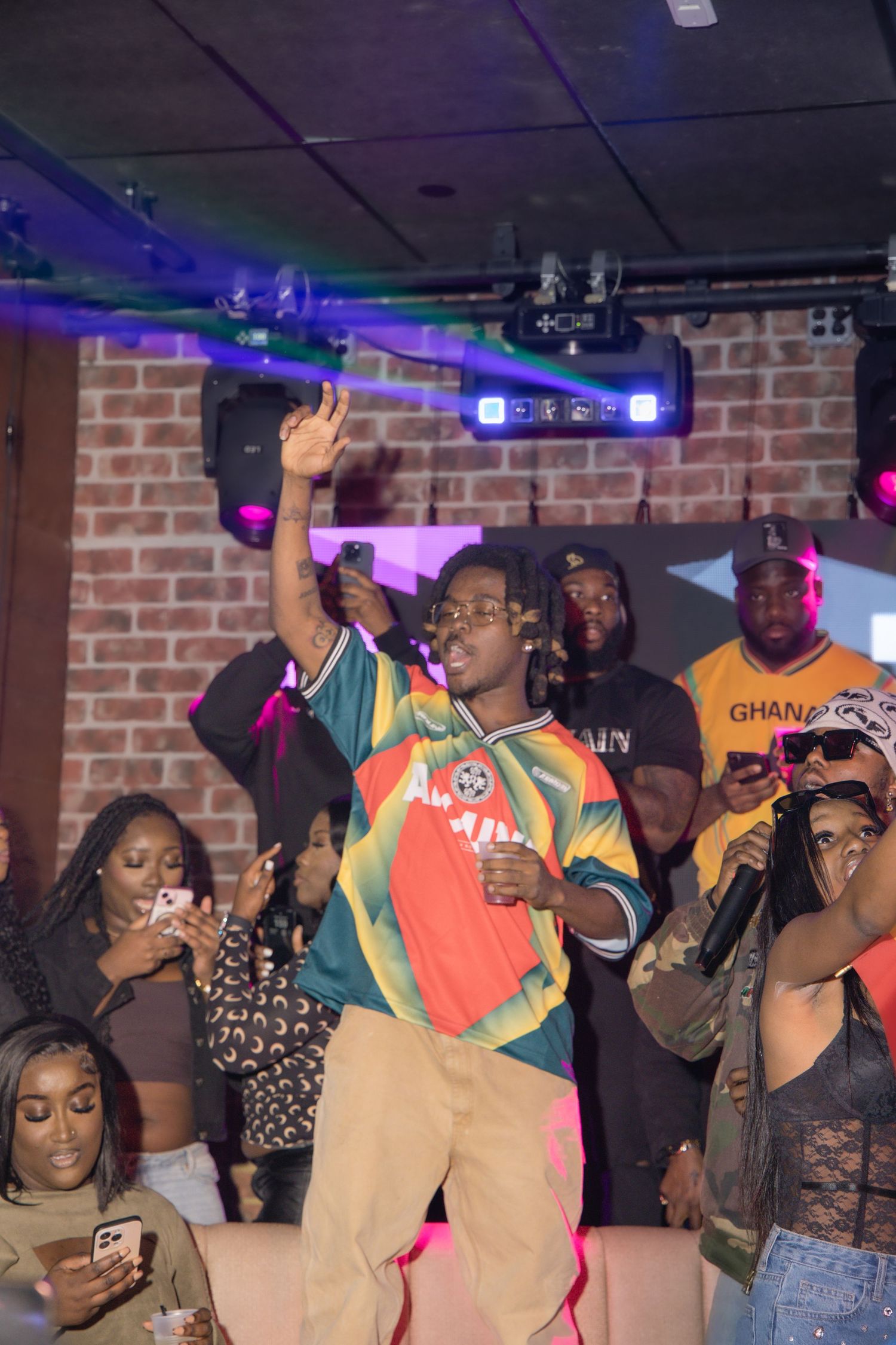 Person in colorful Ghana jersey dancing at a club with purple lighting.
