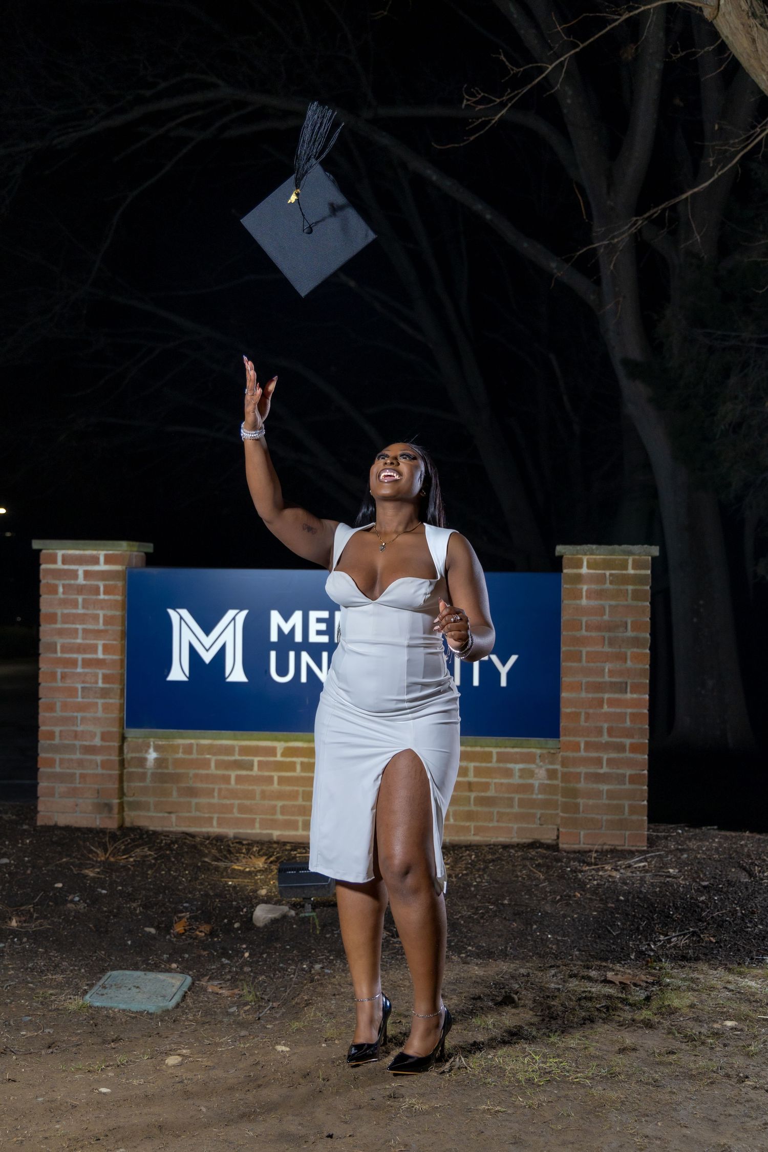 A graduate in white dress throws graduation cap in the air at night in front of a university sign.