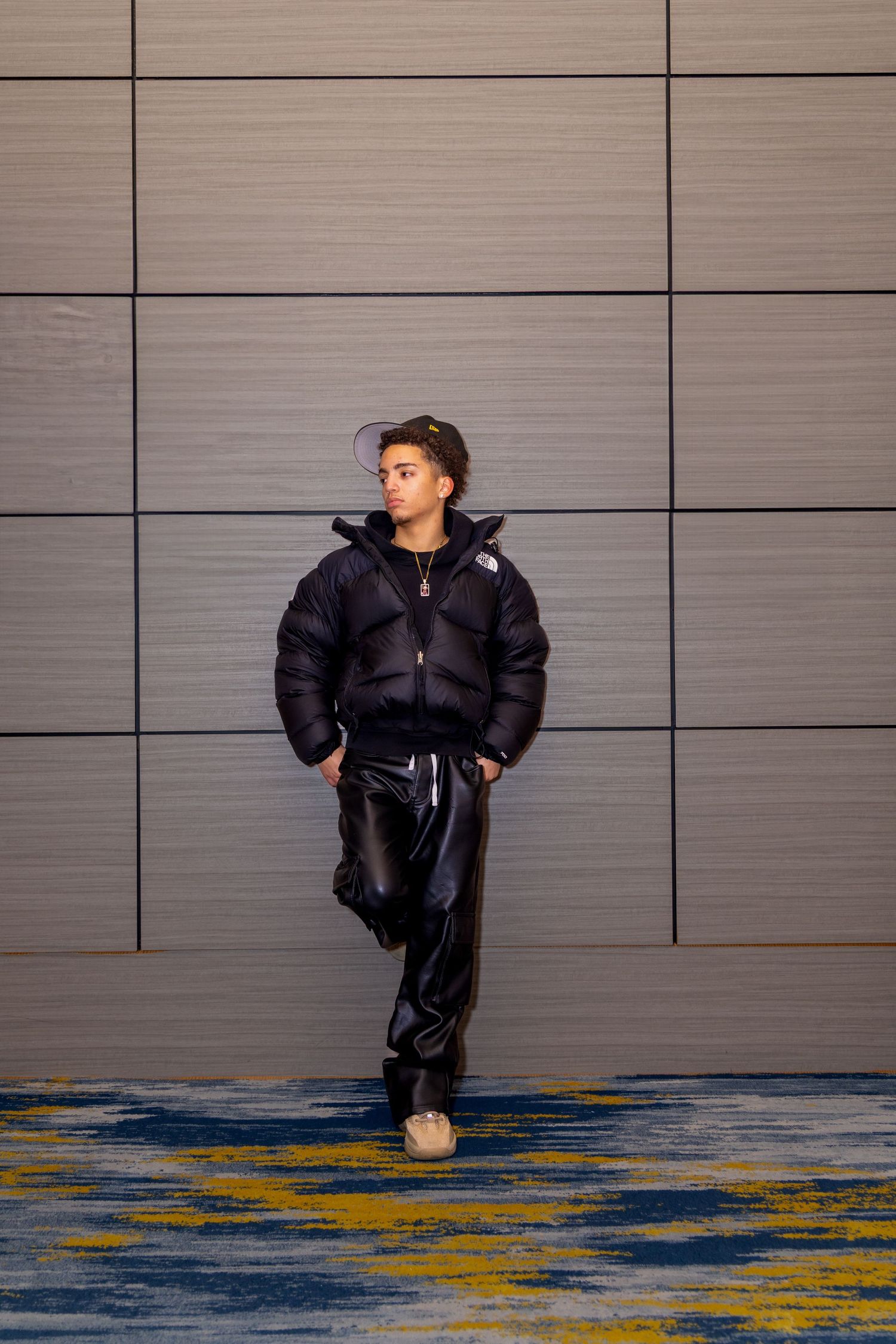 Person in puffy black jacket poses against modern paneled wall with patterned carpet.