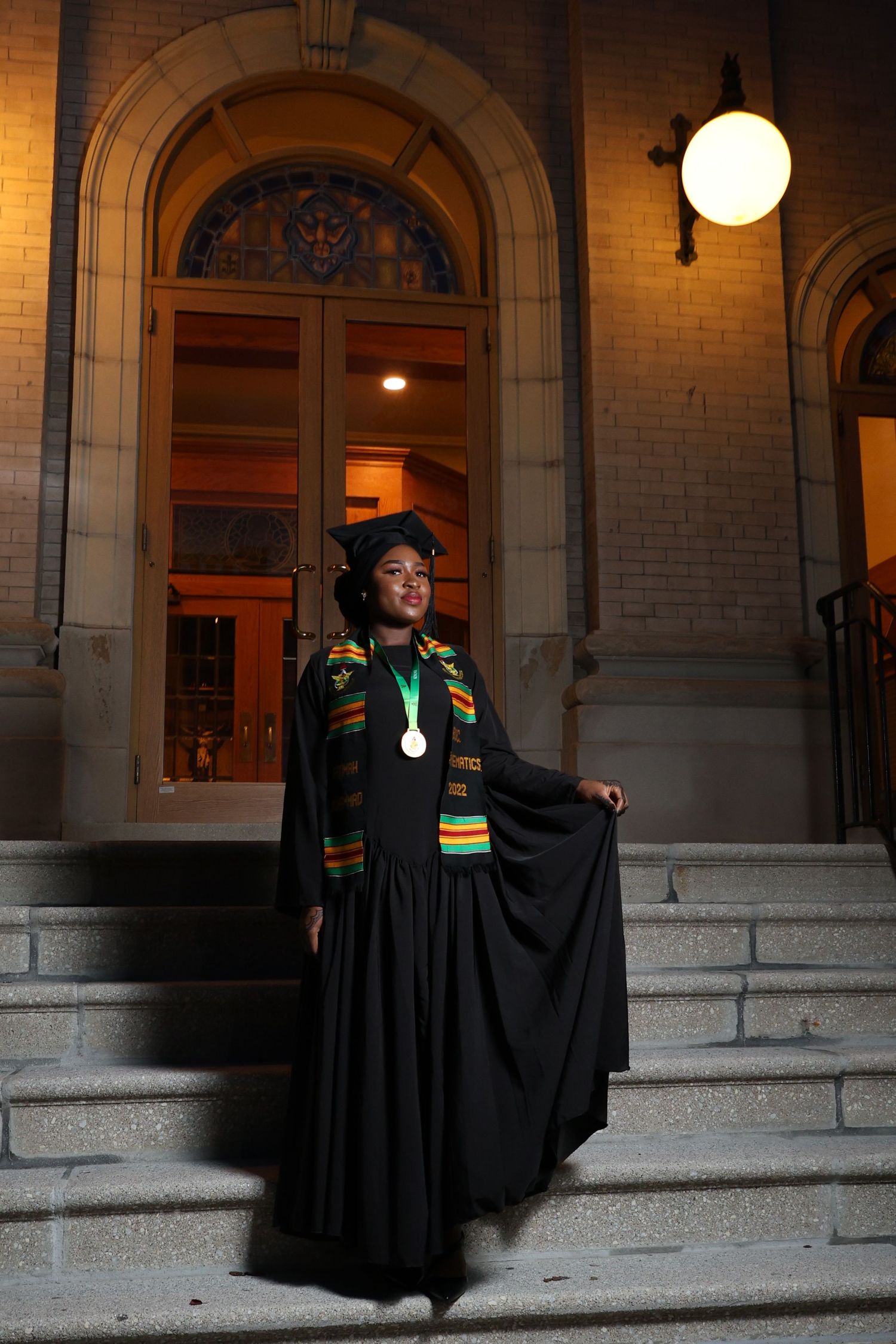 Graduation photo on stone steps featuring academic regalia with colorful stoles and medallions.