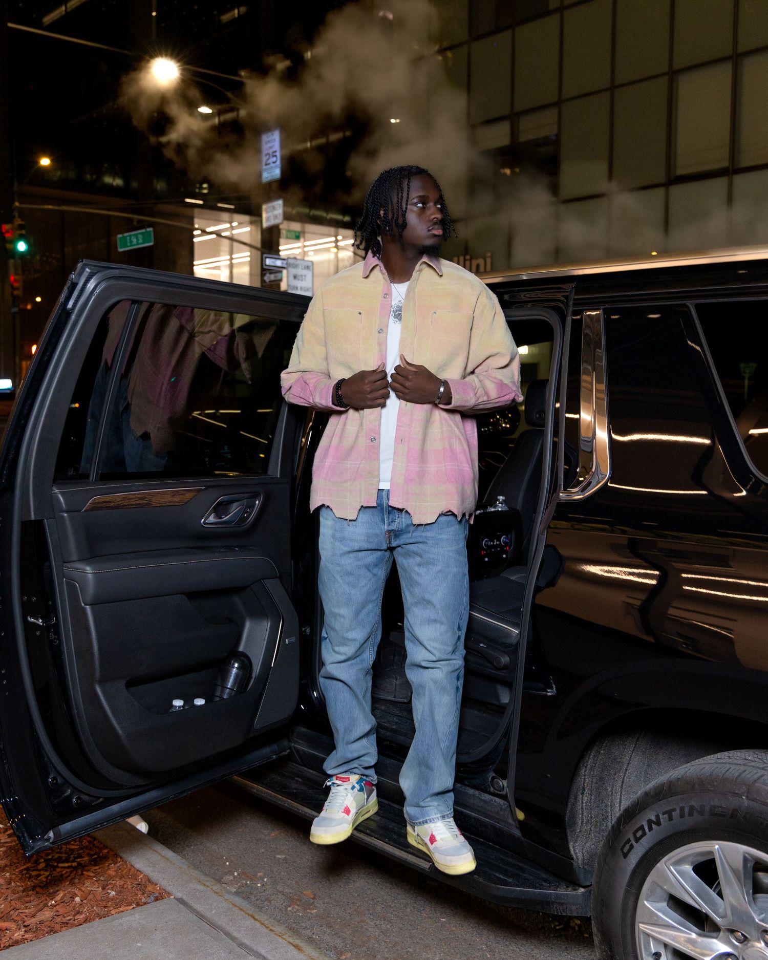 Person in pink button-up shirt and jeans stepping out of black SUV at night.