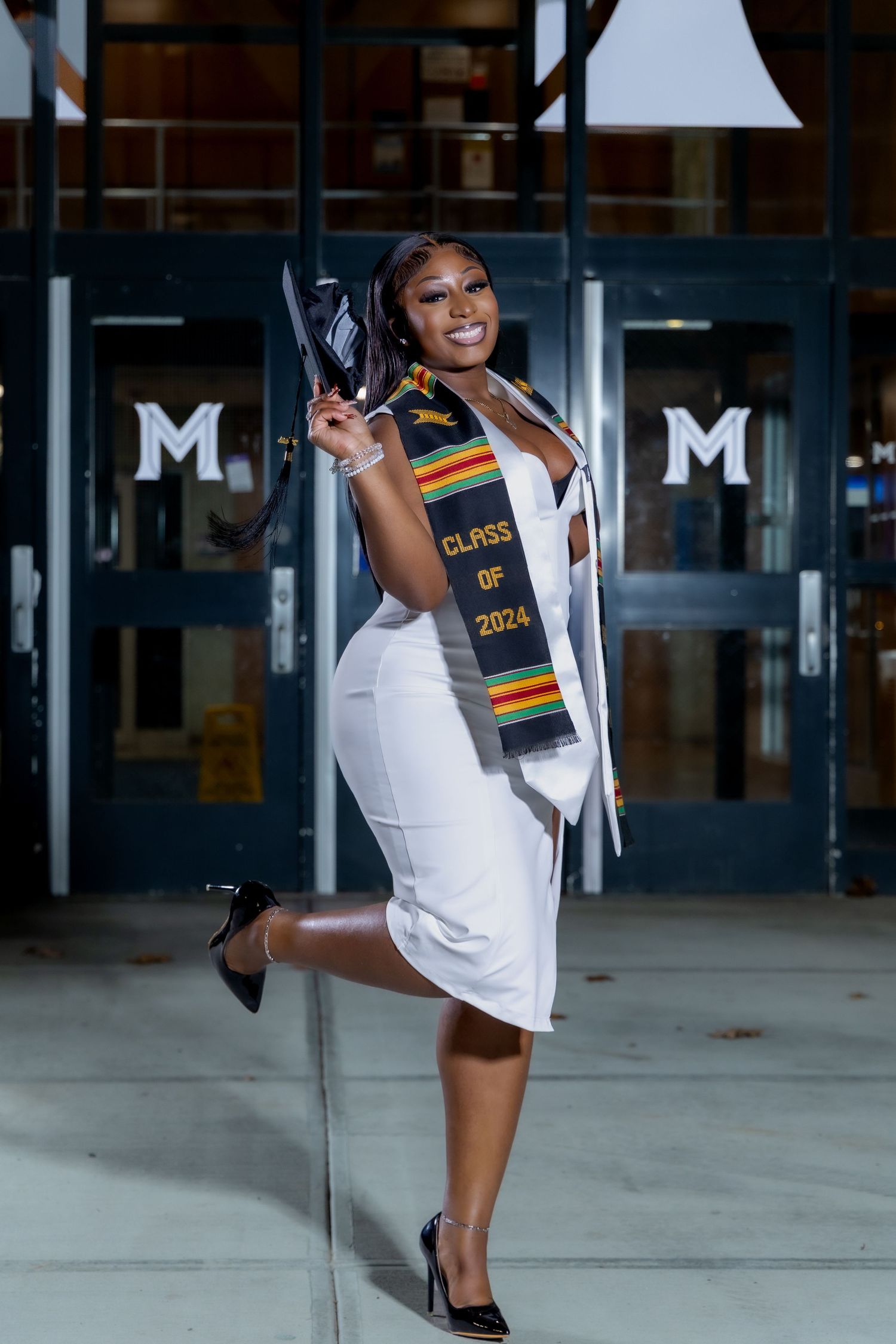 Graduate in white dress and cultural stole poses with diploma outside building.