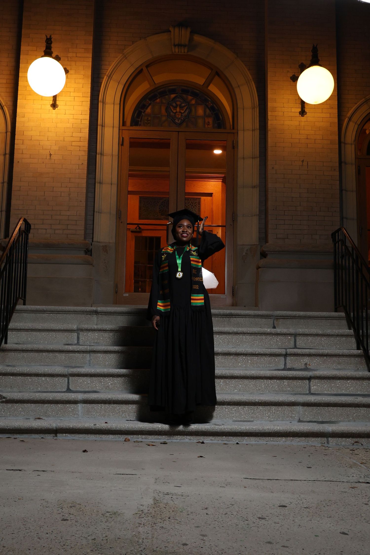A graduate in black graduation gown stands on illuminated steps of a historic building entrance.