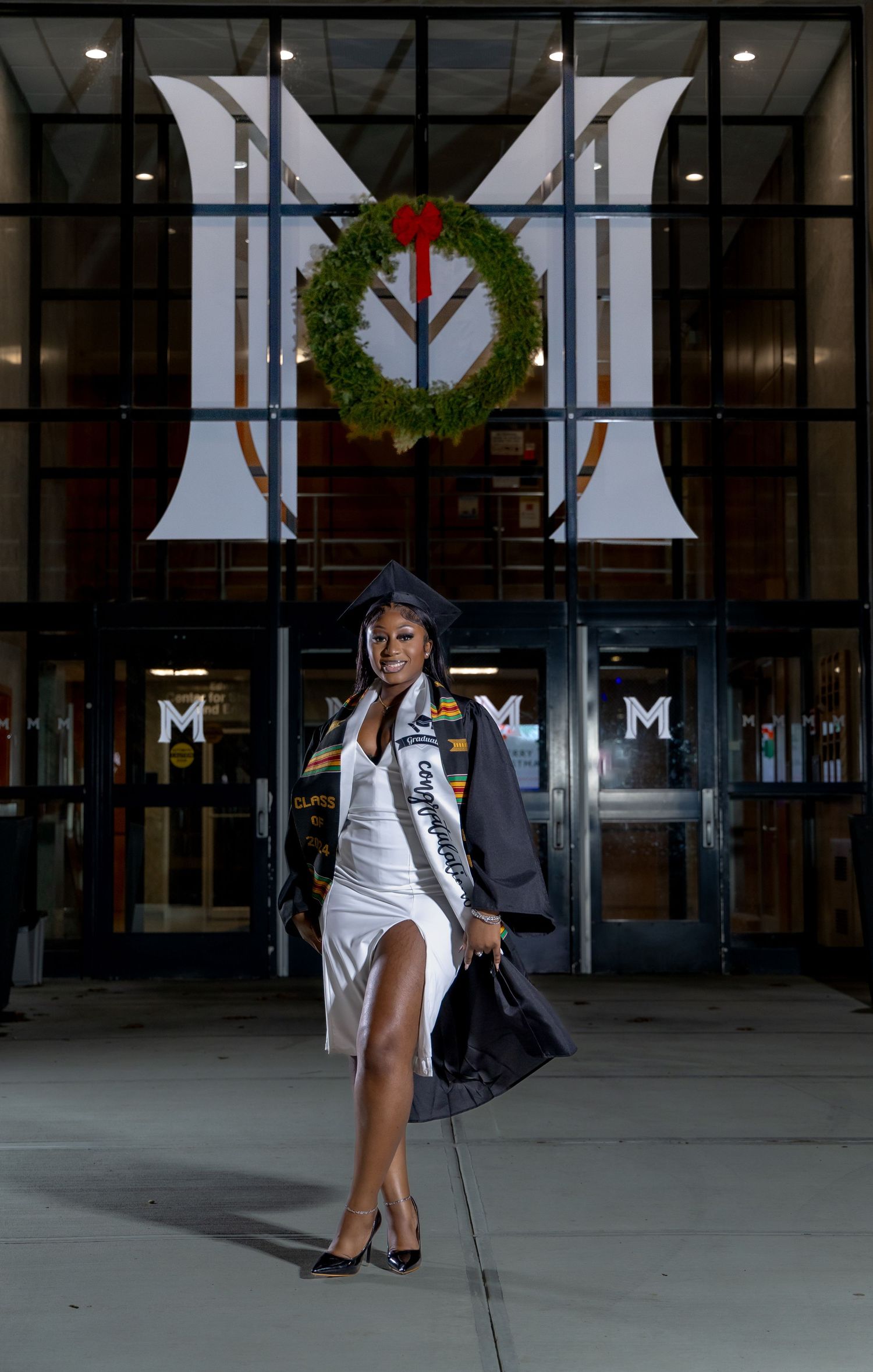 A graduate poses in a white dress and graduation sash in front of a university entrance decorated with a holiday wreath.