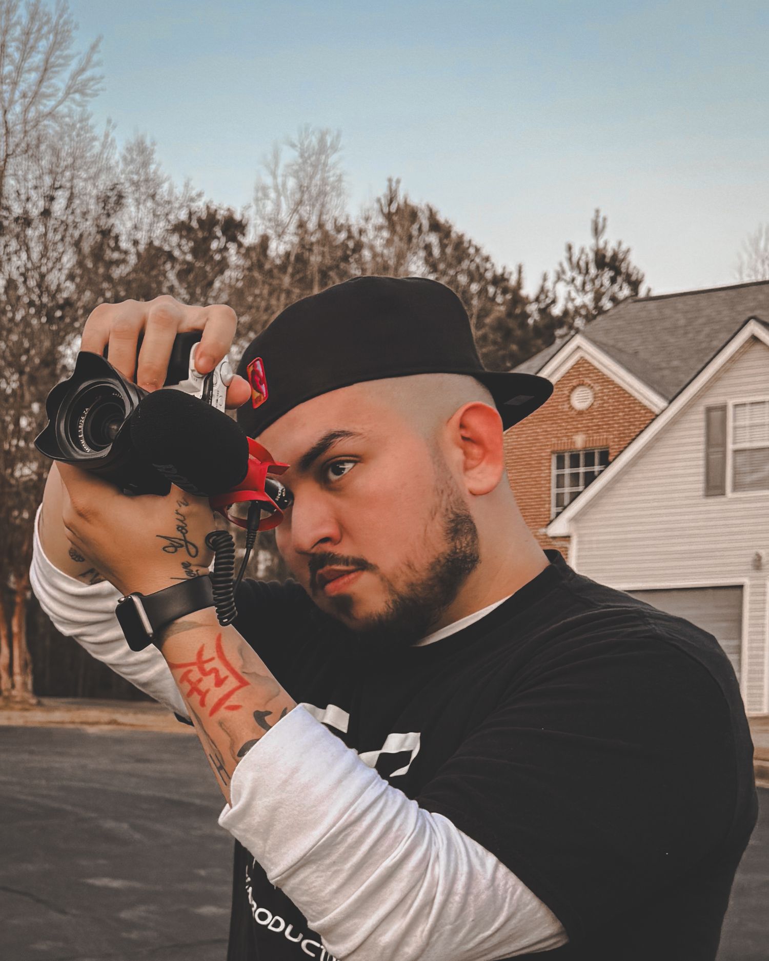 Tattooed man with black hat holding a camera outdoors near a suburban house with bare trees in background.