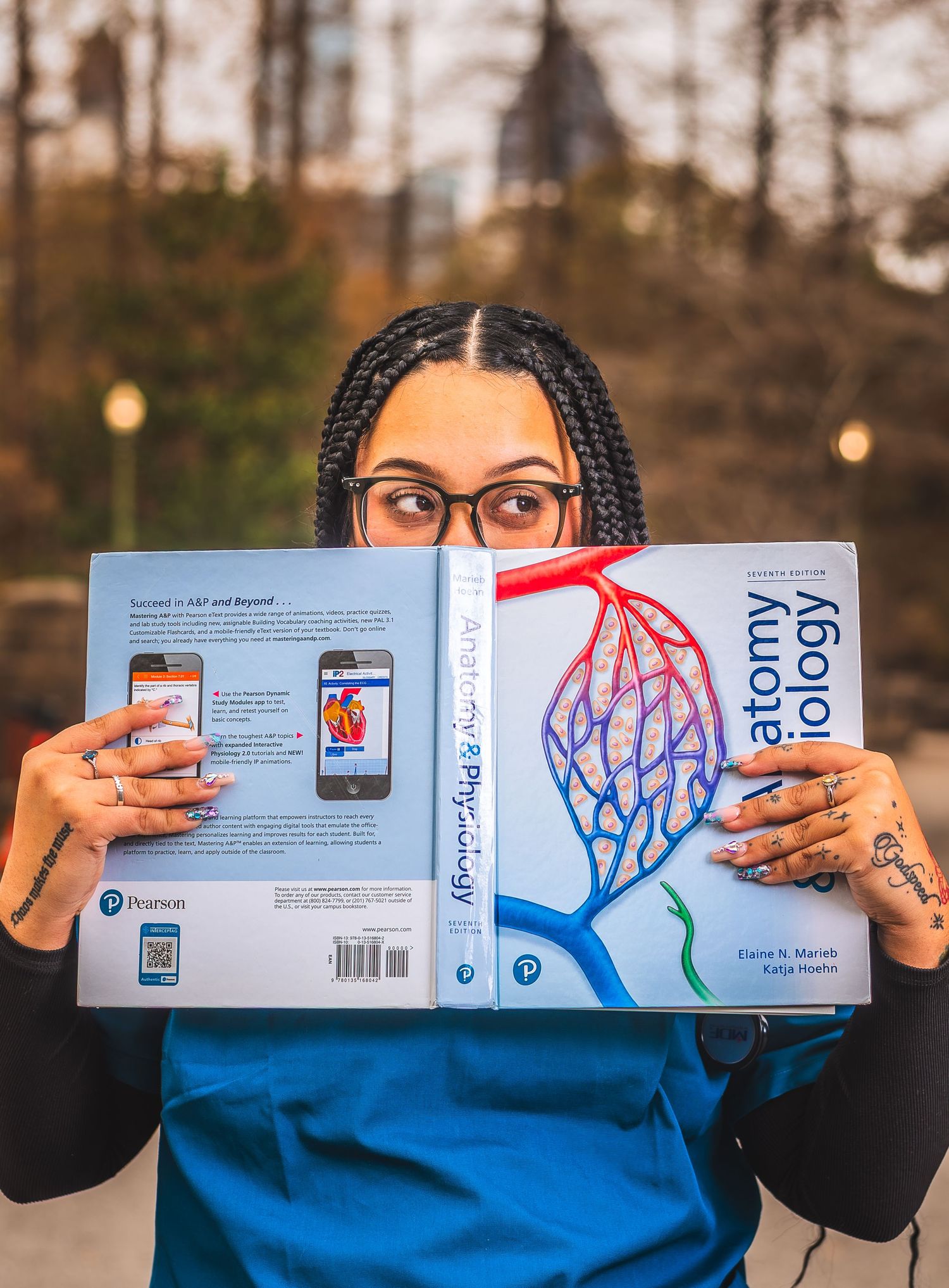 Student holding Anatomy & Physiology Pearson textbook covering her face, wearing scrubs and glasses outdoors.