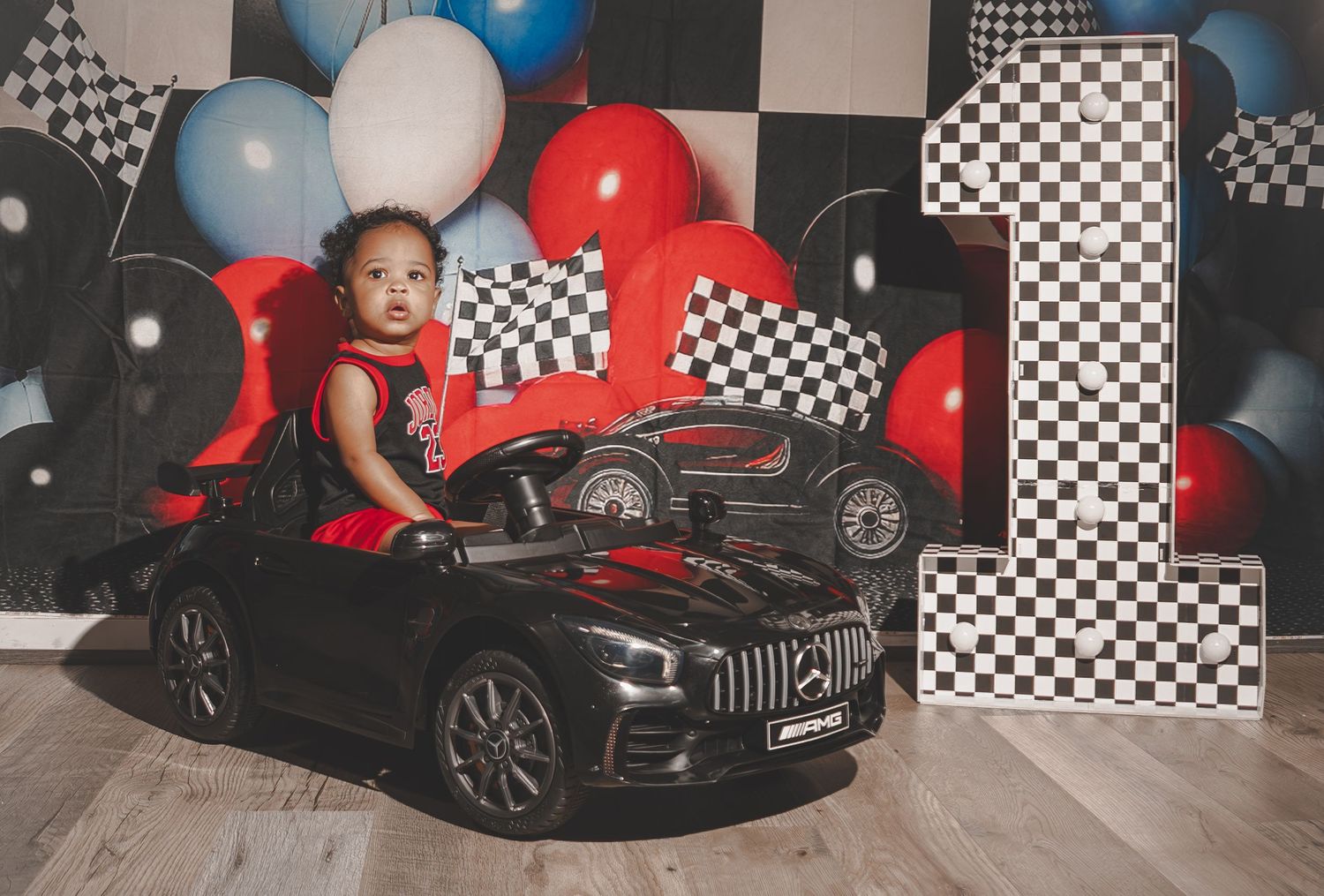 Black luxury car parked in front of racing-themed party backdrop with red and blue balloons and checkered flags.
