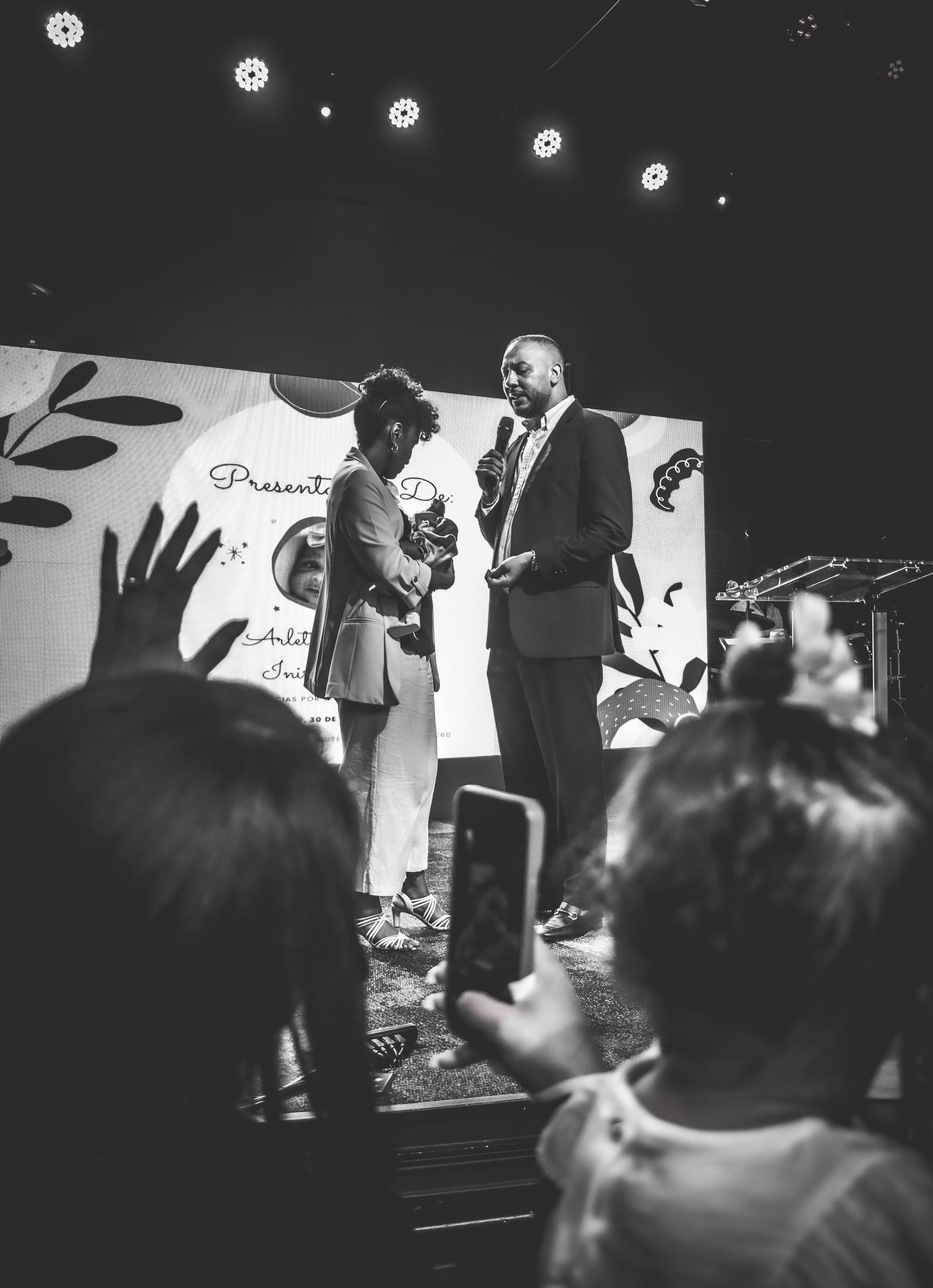 Two speakers share a moment on stage at an event while audience members capture photos on their phones.
