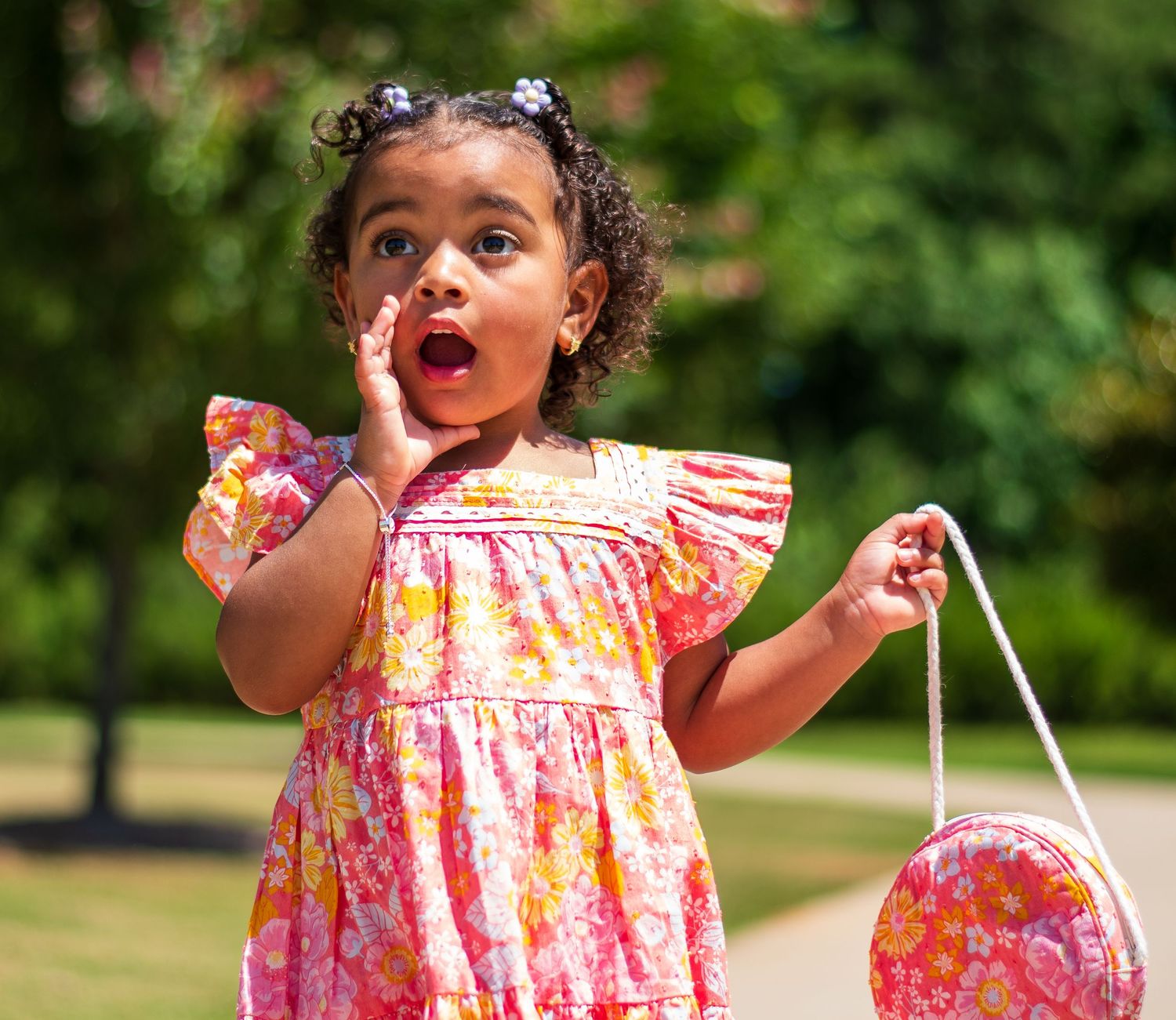 Young girl in floral dress shows surprised expression while holding pink purse outdoors.