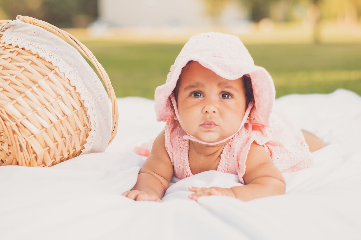 Adorable baby girl in pink bonnet and outfit lying on white blanket next to wicker basket outdoors.
