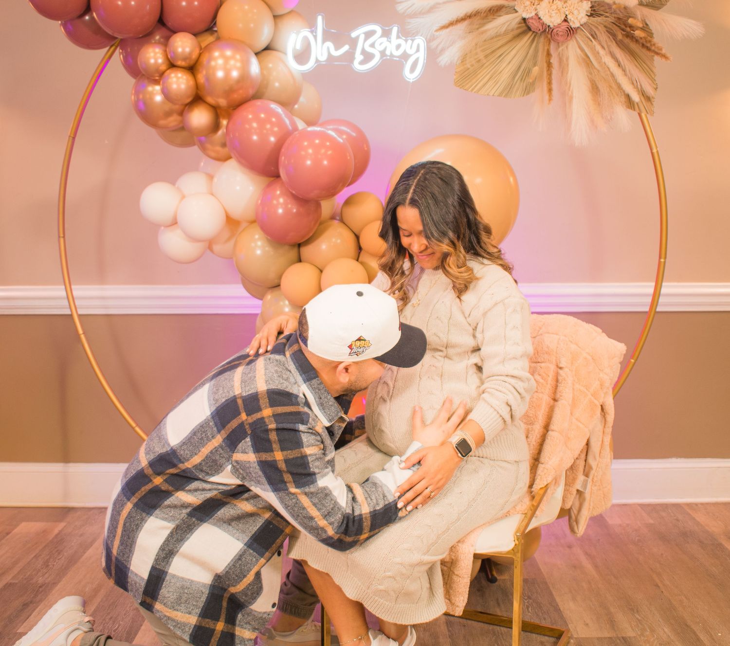 A pregnancy reveal photo with pink and gold balloon decor and the words 'Oh Boy' on the wall.