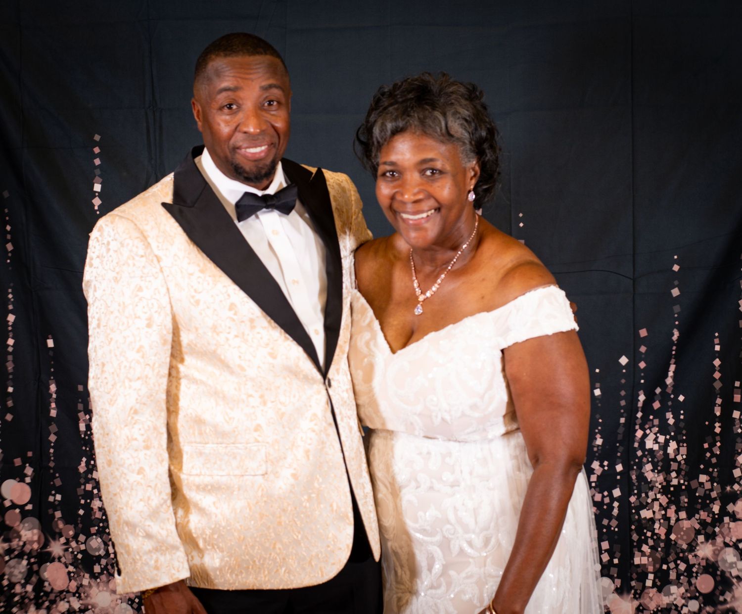 Couple in formal white and black attire posing against sparkly backdrop.