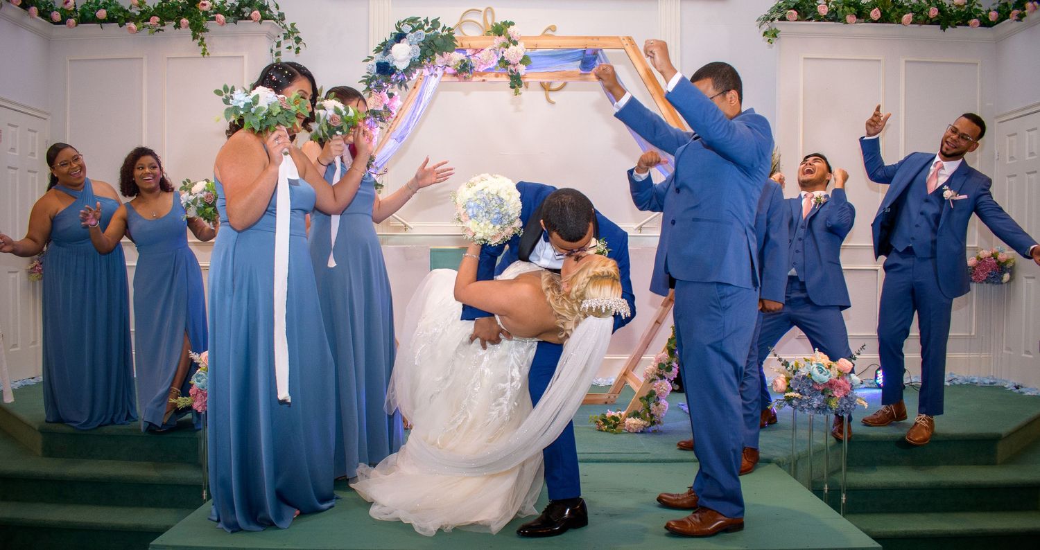 Wedding party in blue attire celebrates with playful poses under a decorated arch.