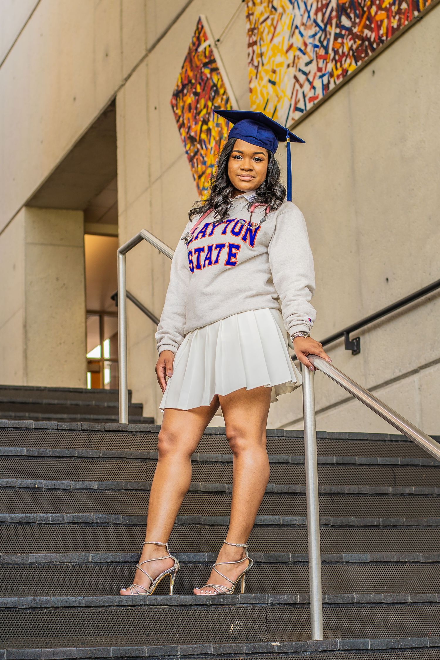 A graduate in Clayton State University attire poses confidently on concrete campus steps.